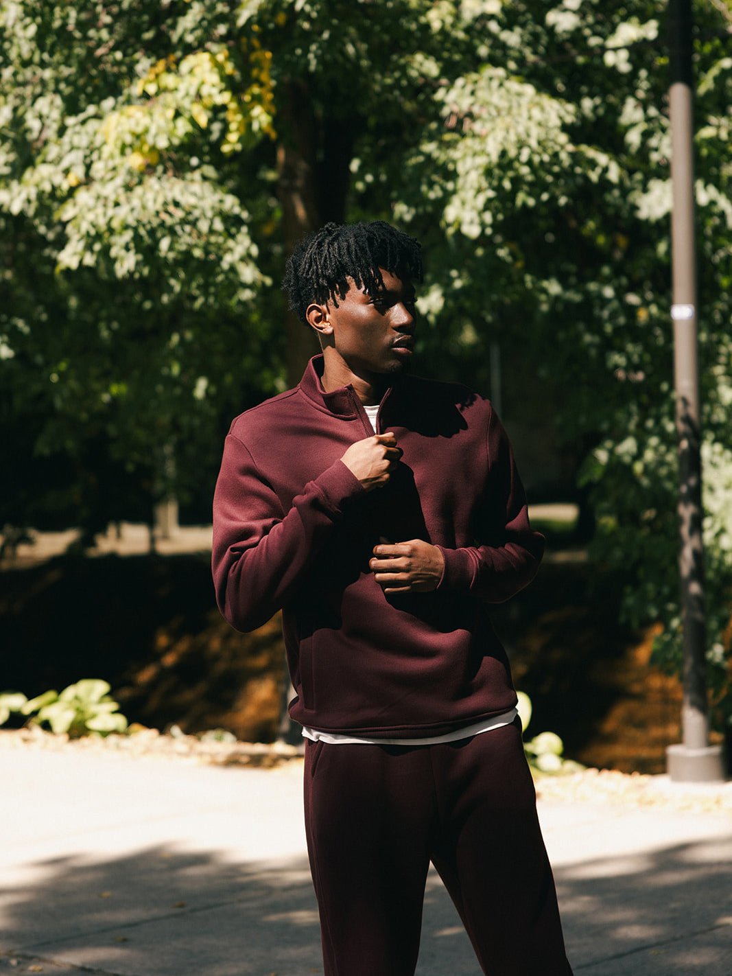 A person is standing outdoors wearing the burgundy Cozy Earth Men's CityScape Quarter Zip with matching pants. They are looking down and touching the collar of their top with one hand. The background includes trees and greenery in a sunlit area. 