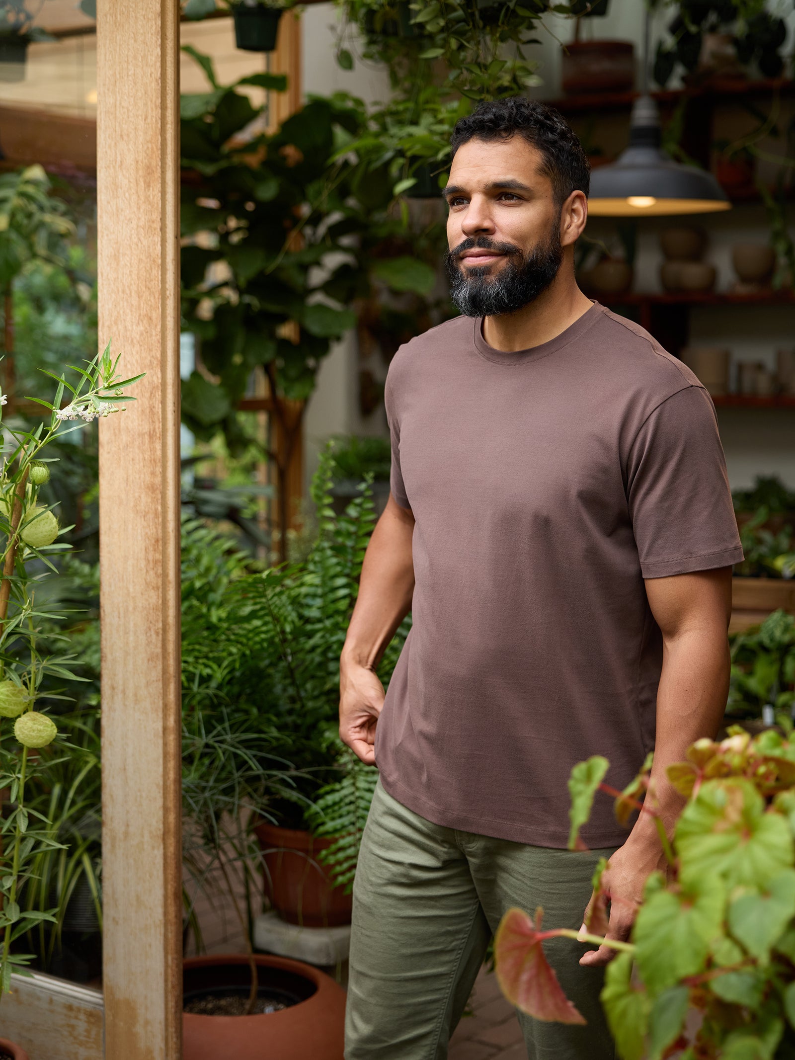 A bearded man wearing a Cozy Earth Men's All Day Tee and green pants stands in a lush plant store, surrounded by various green plants and foliage. He is smiling and looking off to the side, basking in the cozy atmosphere filled with natural light. 