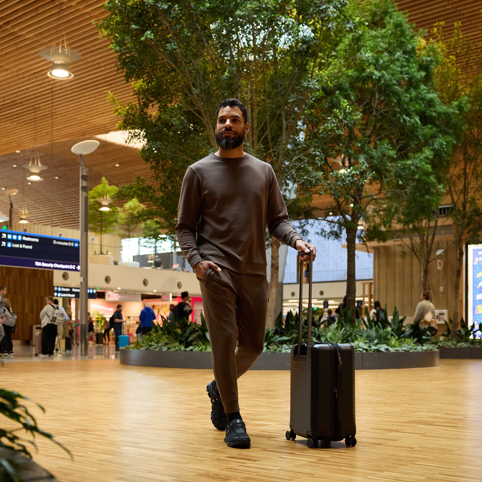 A man with a beard walks through a modern airport terminal while pulling a small black suitcase. He is dressed in Cozy Earth's Men's StretchTech Crewneck, paired with brown tracksuit pants and black shoes. The terminal features wooden floors, green plants, large signs, and travelers in the background. 