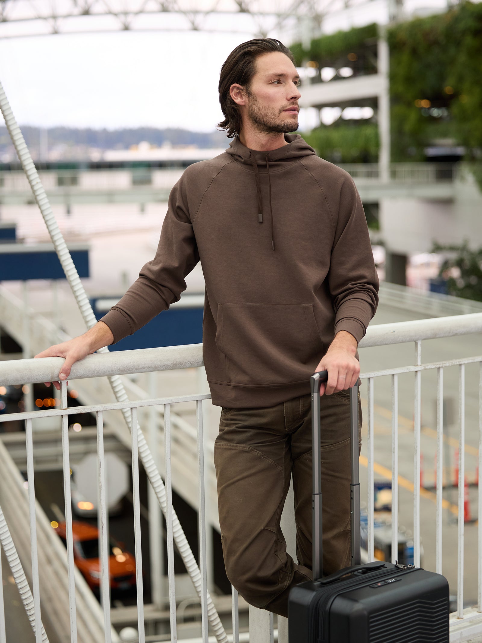 A man in a Cozy Earth Men's StretchTech Hoodie and matching pants stands near a railing at an airport. He is holding the handle of a black wheeled suitcase and looking off into the distance. The background shows airport structures and parked vehicles. 