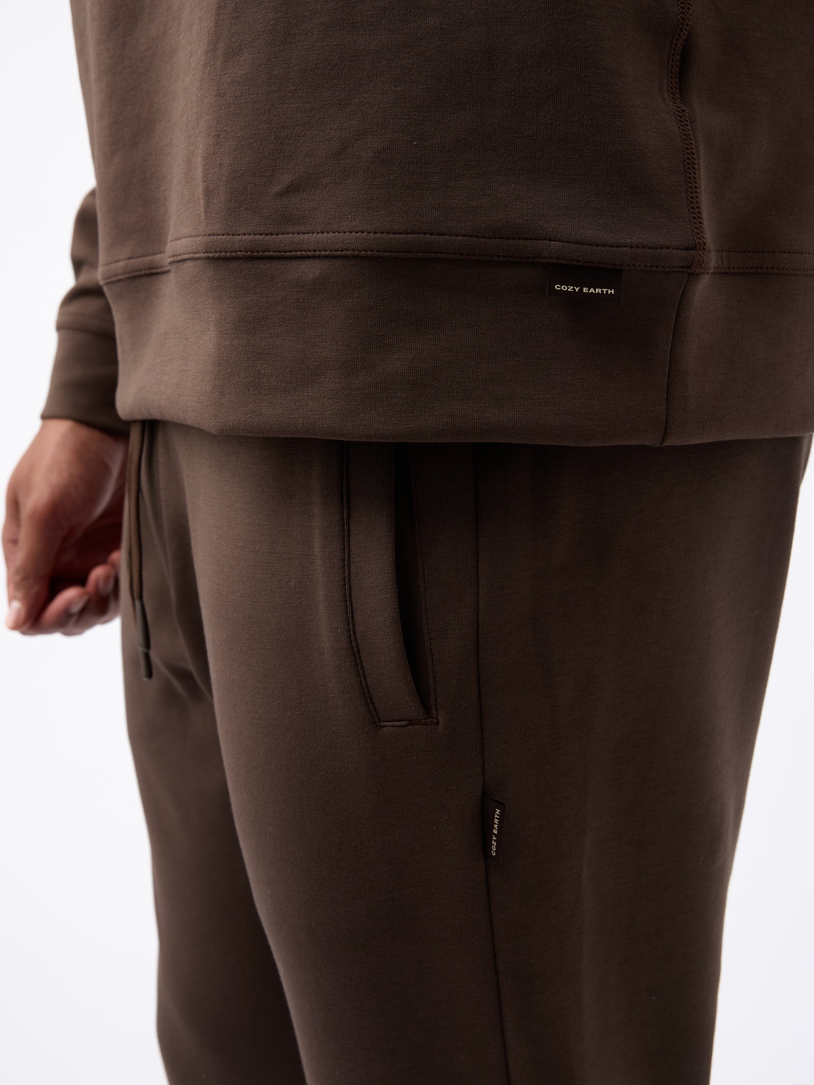 A person is wearing a brown long-sleeve top and matching Men's StretchTech Jogger pants. A small tag on the hem of the top reads "Cozy Earth." The person's left hand is partially visible, hanging by their side. The fabric appears soft and comfortable. 