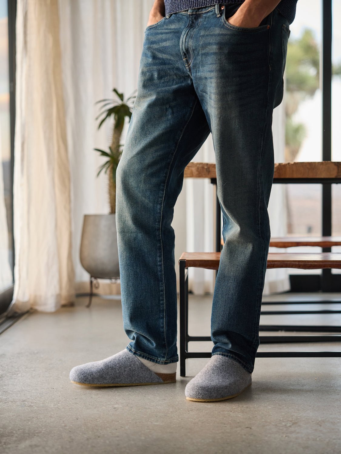 A person in blue jeans and Cozy Earth's gray Lakehouse Clogs stands in a sunlit room with hands in pockets. A wooden bench, a potted plant, and sheer curtains are visible in the background. 