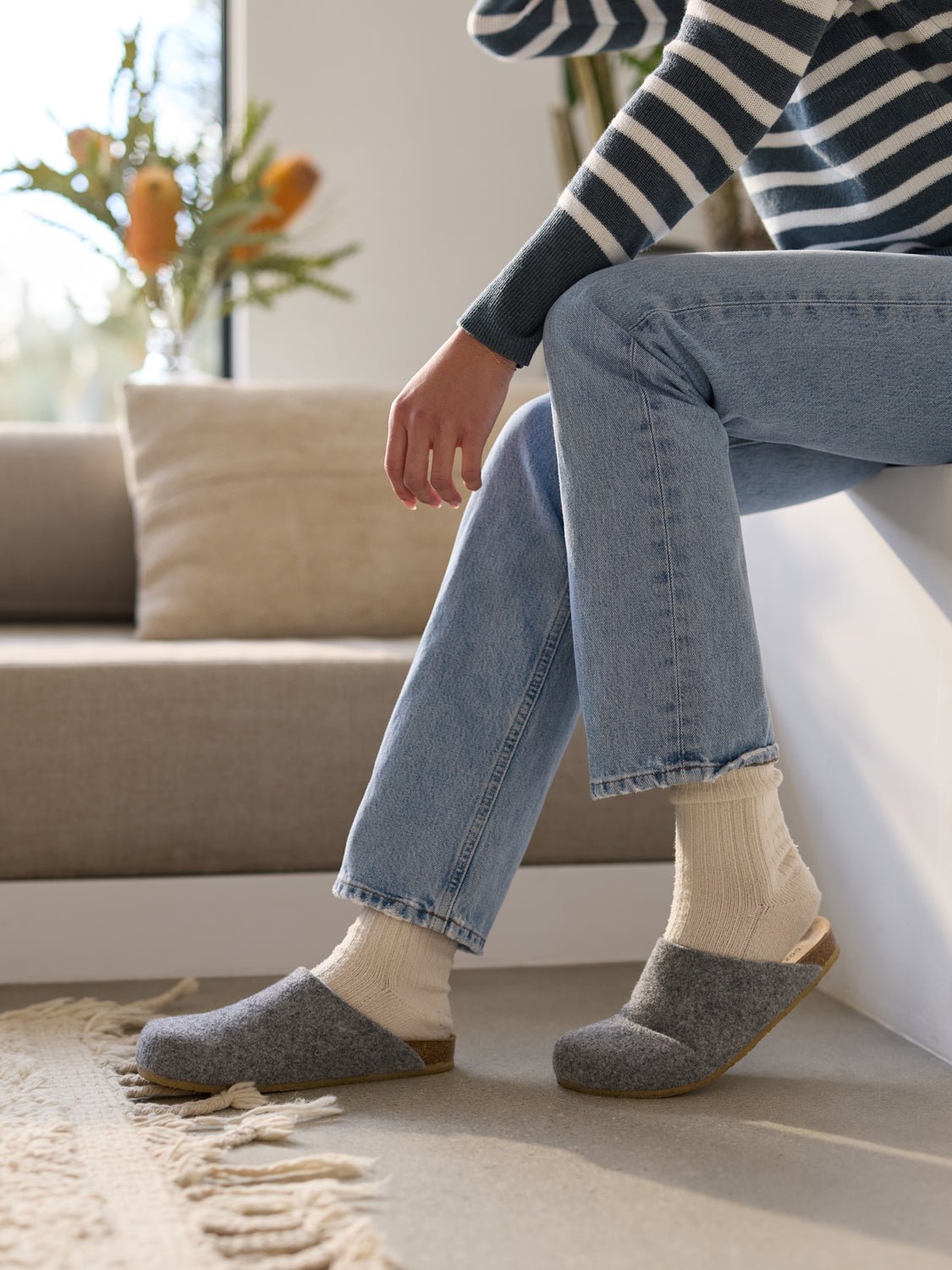 A person in a striped sweater, light jeans, and Cozy Earth's Lakehouse Clog sits on a white bench with one foot crossed over the knee. A beige rug is underfoot, and flowers adorn the bright window in the background. 