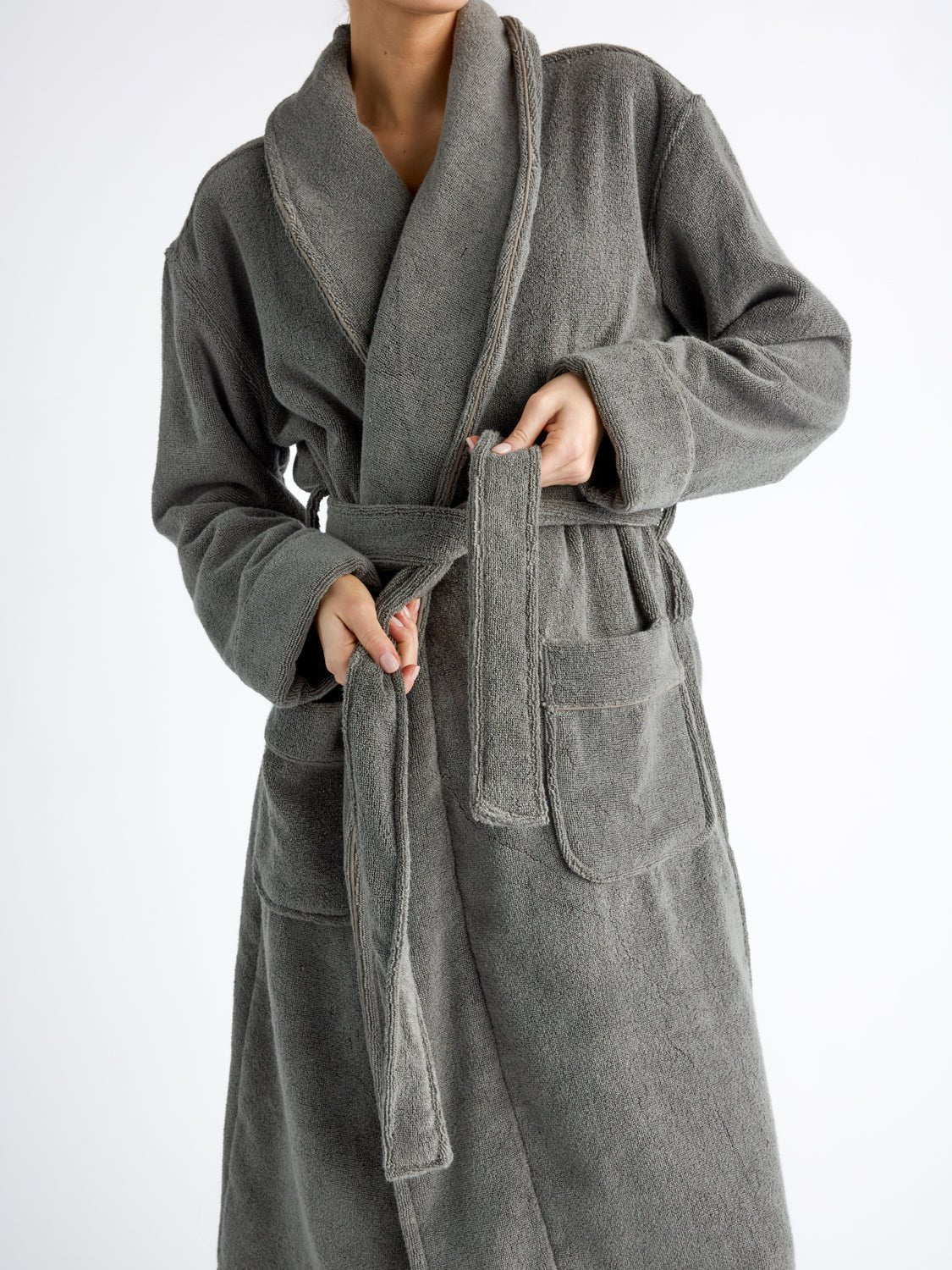 A close up of a woman from the shoulders to her knees tying the belt of a Cozy Earth Luxe Bath Robe in Charcoal. 