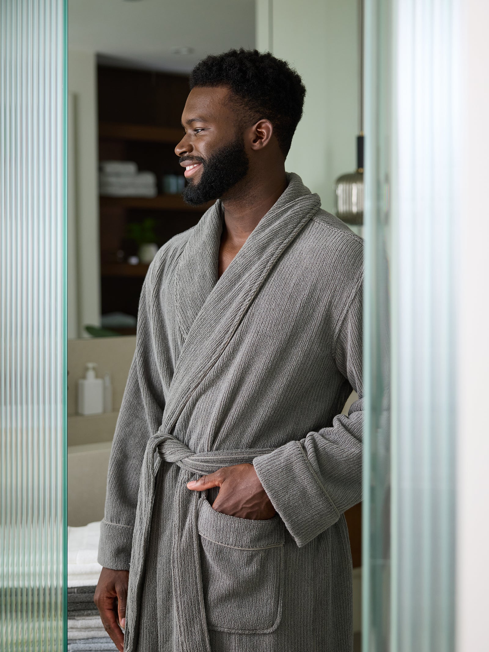 A man stands in a doorway, wearing a Cozy Earth Ribbed Terry Bath Robe and smiling while looking to the side. The background shows a neatly organized room with shelves and folded towels. 