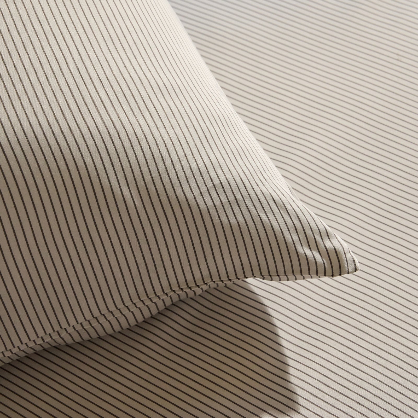 A close-up view showcases the Bamboo Fitted Sheet by Cozy Earth alongside a pillow, both displaying thin, evenly spaced vertical stripes in shades of gray and white. The texture and shadow of the fabric suggest an exceptionally soft and comfortable material. 