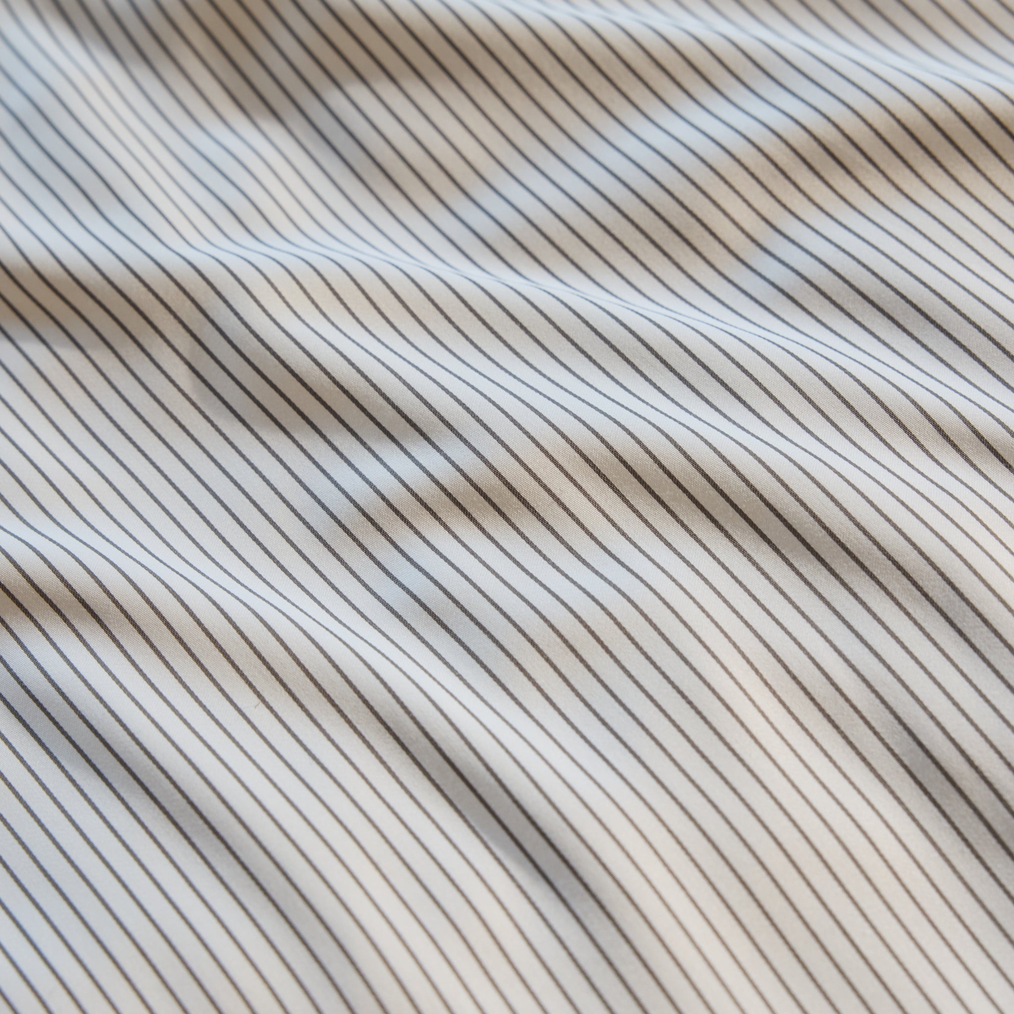 Close-up of the Bamboo Sheet Set by Cozy Earth, featuring a white fabric with thin black vertical stripes. The slightly wavy fabric creates gentle shadows and highlights, imparting a sense of texture and movement to the image. |Color:Charcoal Pinstripe