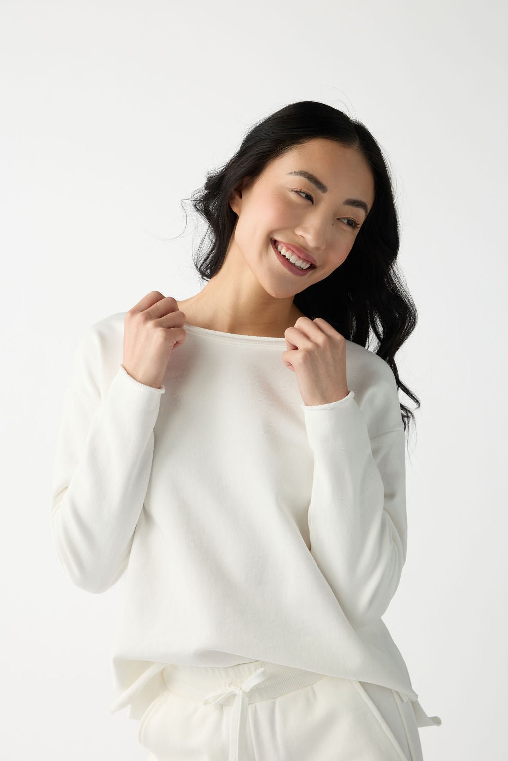 A person with long dark hair smiles while gently pulling at the neckline of a white Women's Chelsea Set from Cozy Earth, featuring matching white pants against a plain, light-colored background. 