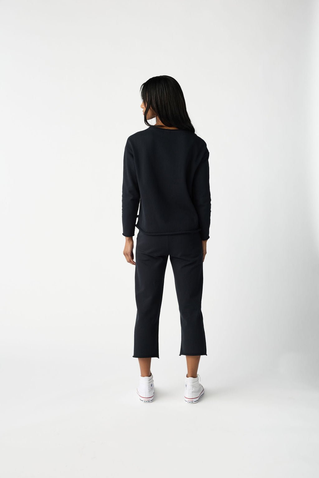 A person with long dark hair stands facing away, wearing Cozy Earth's Women's Chelsea Set in black—a sweater and cropped pants paired with white sneakers. The background is a plain white studio setting. 