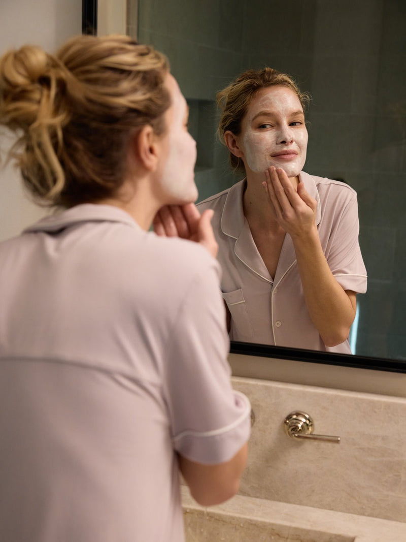 Wearing light-colored pajamas, a person stands before a bathroom mirror, applying Cozy Earth's Clay Mask. They gaze at their reflection while evenly spreading the mask over their cheeks and forehead.