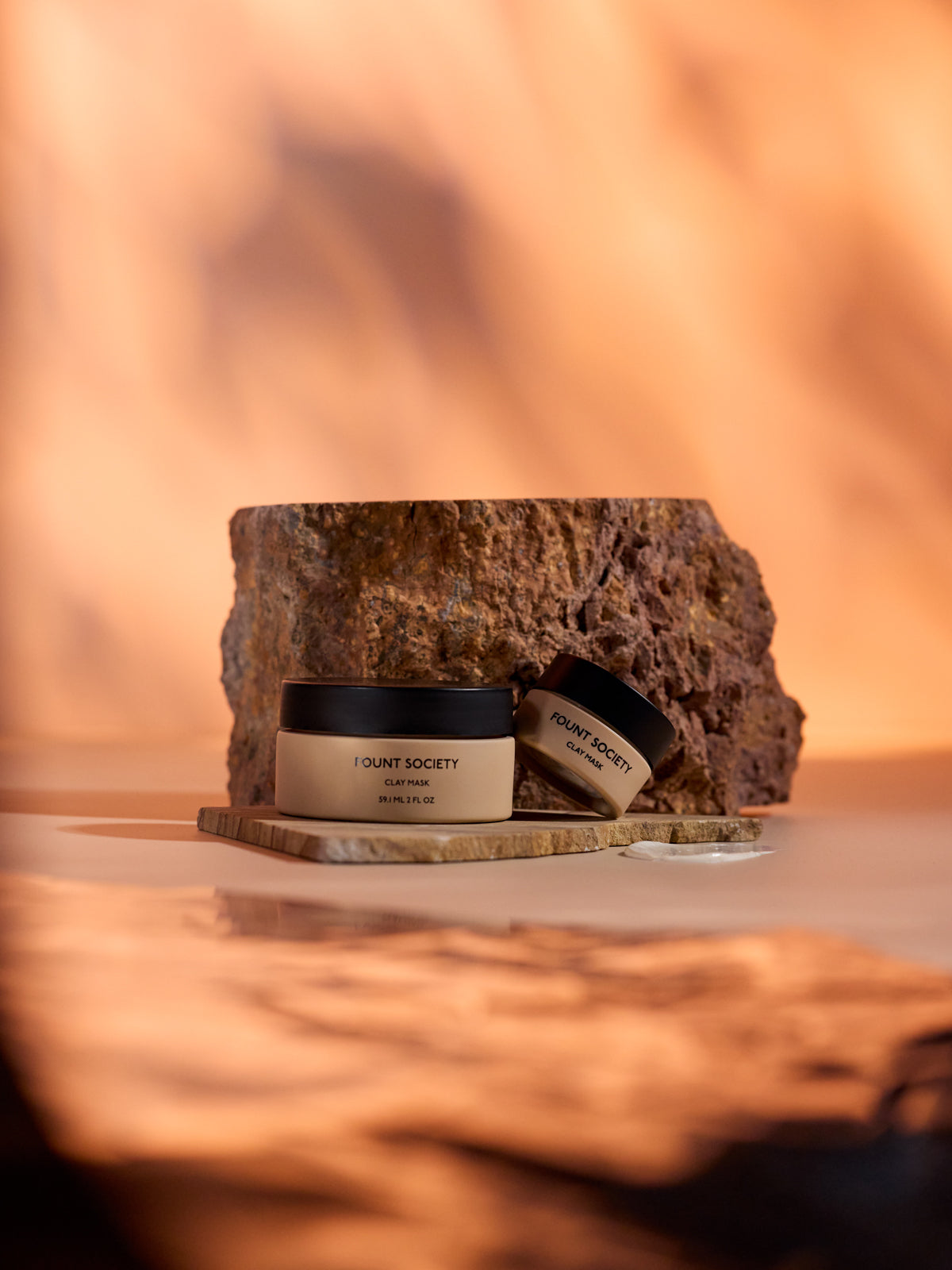 Two Cozy Earth Clay Mask jars sit on a wooden slab against a textured brown rock, with a warm, blurred orange background creating an earthy ambiance. 