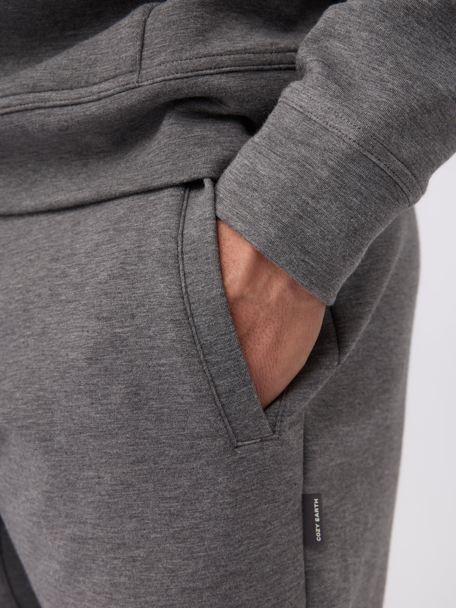 A person sporting the Men's StretchTech Jogger by Cozy Earth in gray, casually placed their right hand in the pants pocket. The fabric looks soft and comfortable, accentuated by a small label on the side of the joggers. 