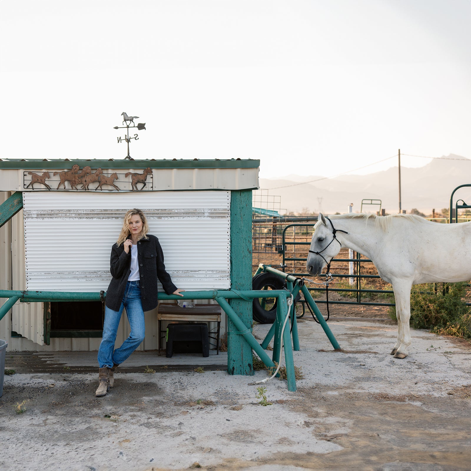 A person wearing a Cozy Earth Women's Boucle Shacket and blue jeans stands outdoors next to a small structure with a weather vane on top, featuring horse decorations. A white horse is tethered to a post near the person. The background includes fencing and distant mountains. 