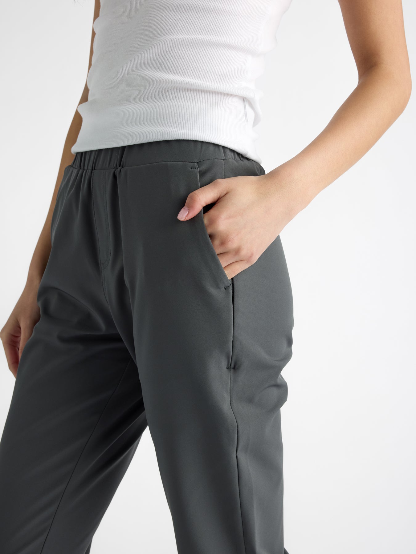 A person models the Women's Always Cropped Pant by Cozy Earth, wearing a white sleeveless top and dark gray pants against a white background, with one hand in their pocket and their head not visible. 