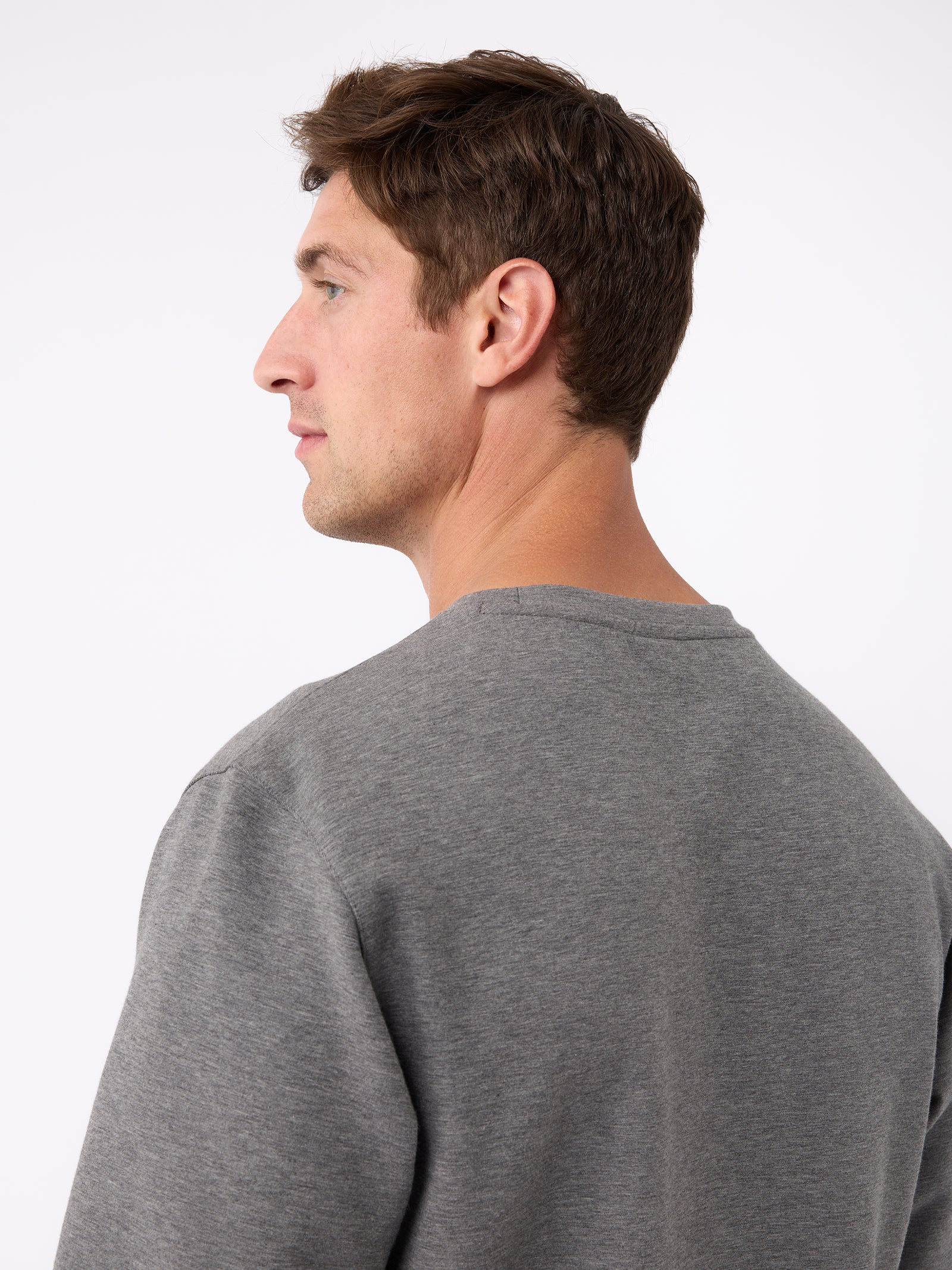 A person with short brown hair is shown from the side, wearing a gray Men's StretchTech Crewneck by Cozy Earth. The background is plain white. 