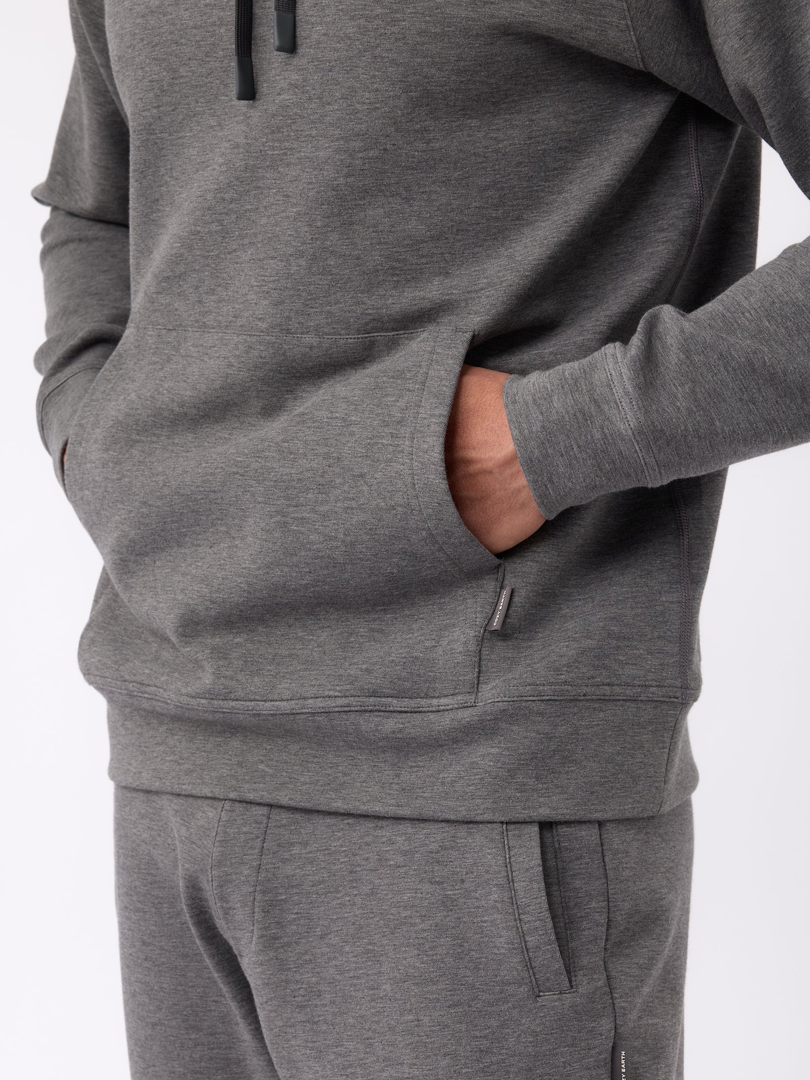 A person stands with one hand in the pocket of a Cozy Earth Men's StretchTech Hoodie, which is gray and part of a matching tracksuit. The background is a plain, light color. 