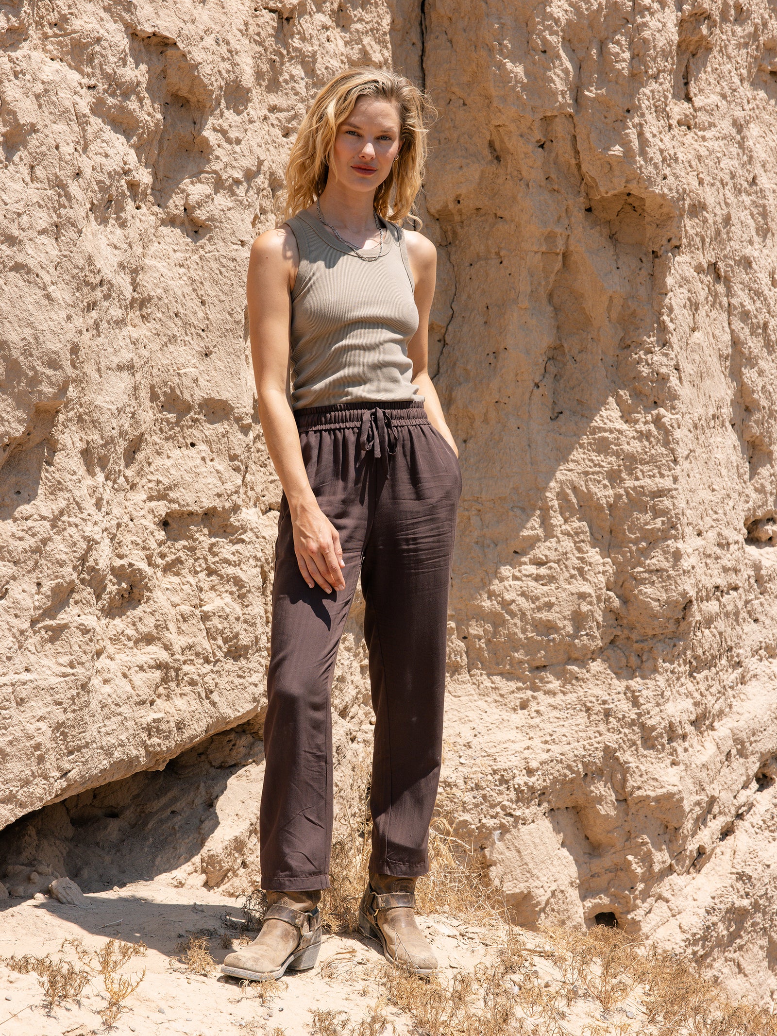 A woman with shoulder-length blonde hair stands against a rocky, sunlit background. She is wearing a sleeveless beige top, Cozy Earth's Women's Sunset Cropped Pant in brown, and brown ankle boots. Her hands are in her pockets as she looks confidently at the camera. 