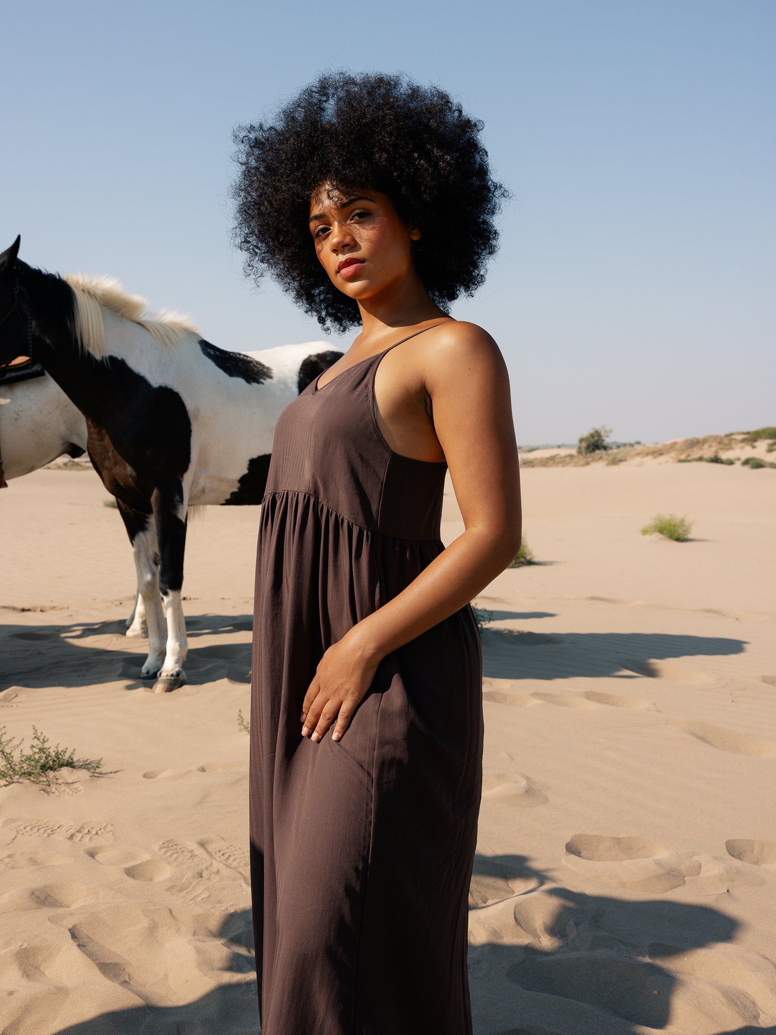 A woman with curly hair wears a loose, dark Cozy Earth Women's Sunset Gathered Dress, standing in a sandy desert landscape. A black and white horse stands in the background under a clear blue sky. 