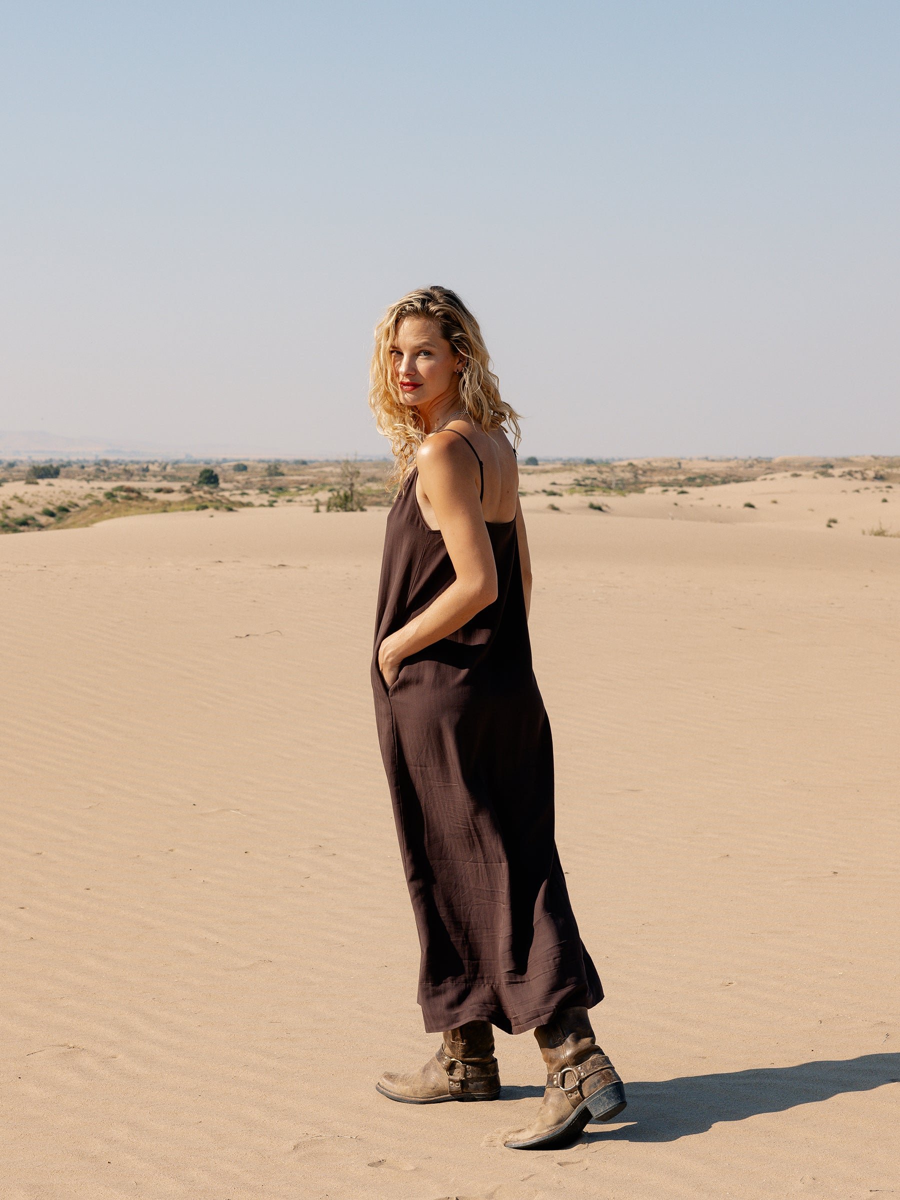 A woman with curly blonde hair, wearing a loose Women’s Sunset Slip Dress from Cozy Earth and brown boots, stands in a sandy desert landscape. She looks over her shoulder at the camera with a slight smile. The sky is clear and blue. 