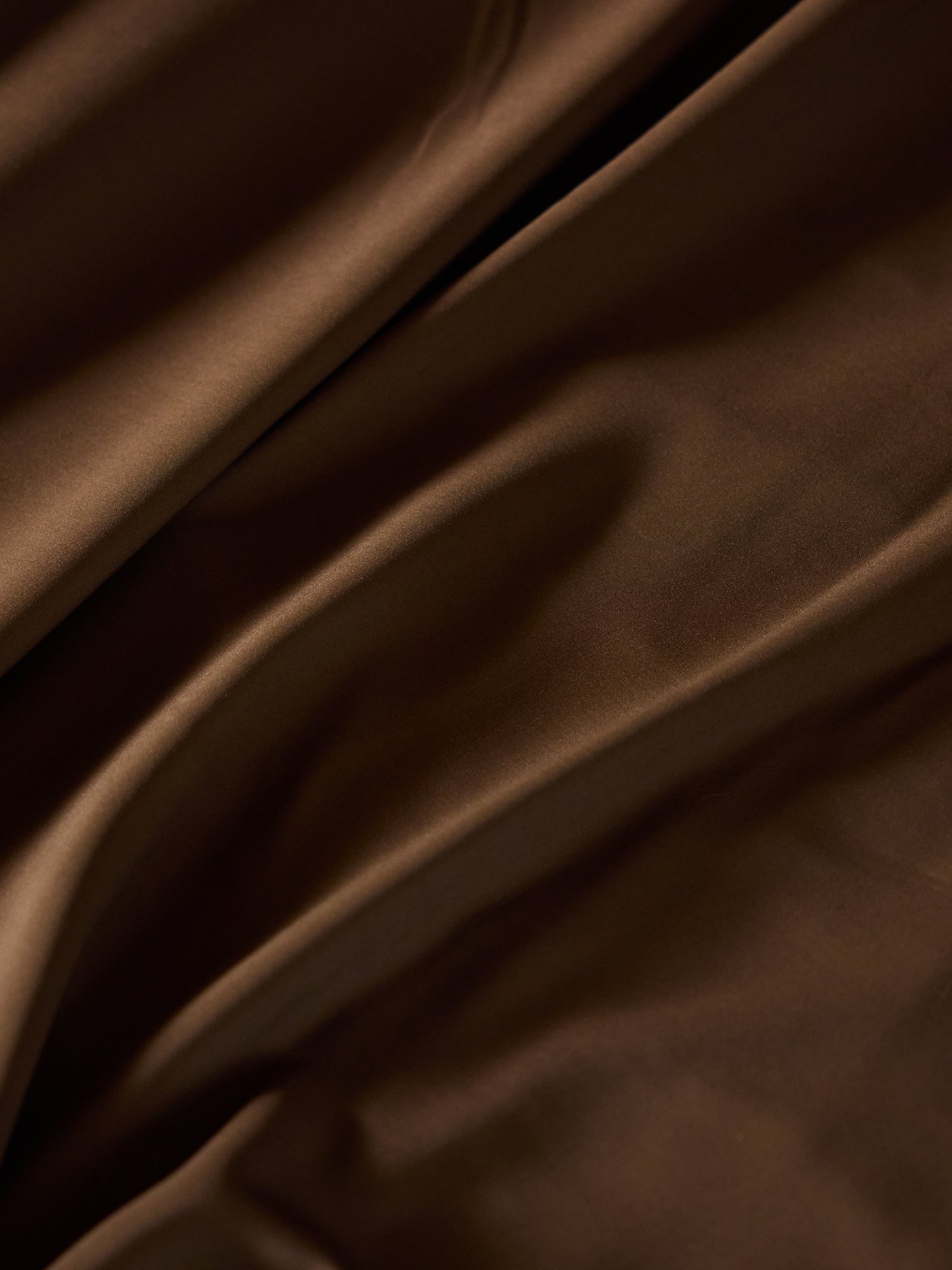 A close-up image of the rich, brown fabric of the Bamboo Fitted Sheet by Cozy Earth shows soft folds and creases. The texture appears smooth and silky, with subtle light reflections creating darker and lighter areas across the sheet. 