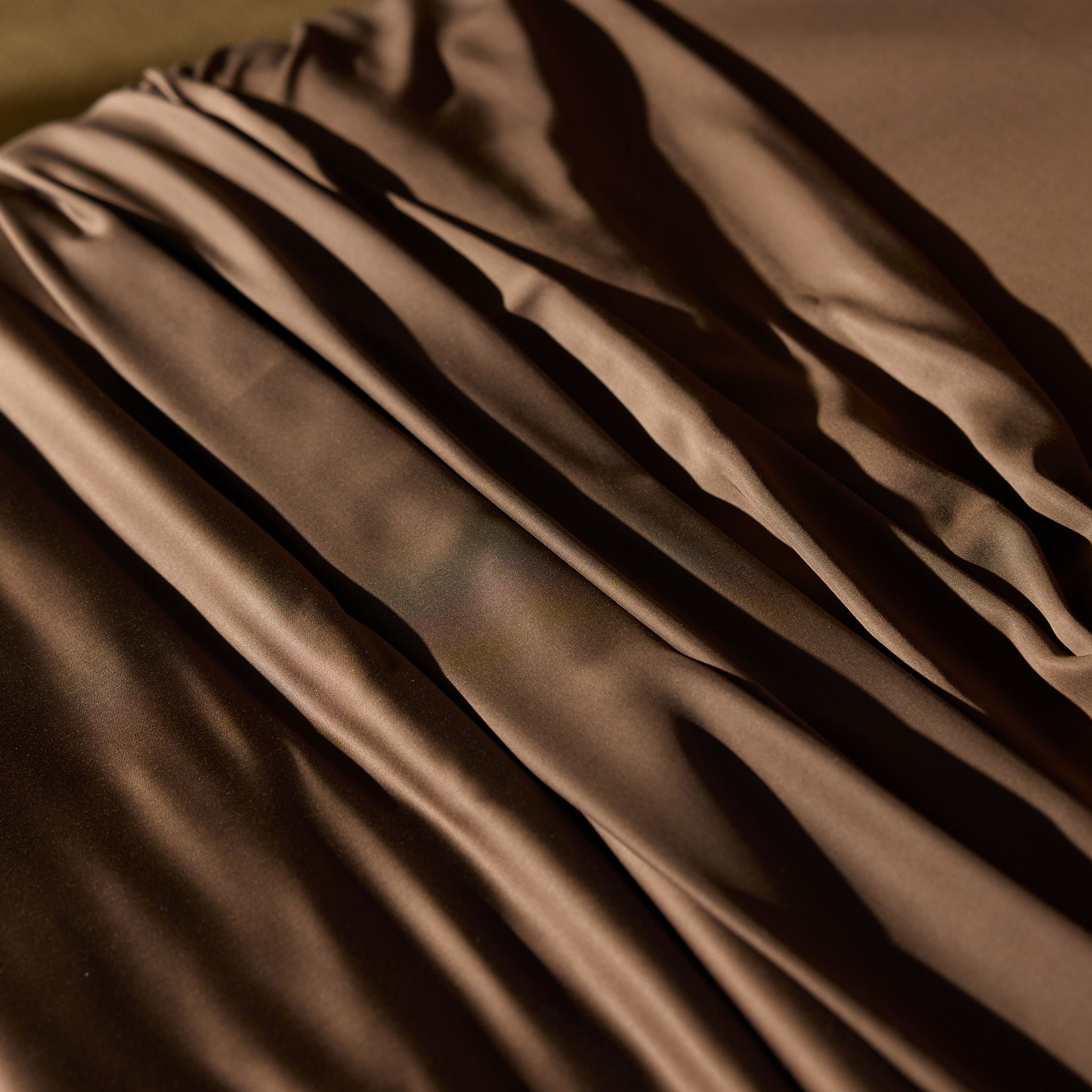 Close-up image of gathered and draped Bamboo Sheet Set by Cozy Earth in a rich brown color, creating soft, flowing folds with highlights and shadow play. The texture appears smooth and slightly shiny, suggesting a luxurious and elegant material. 