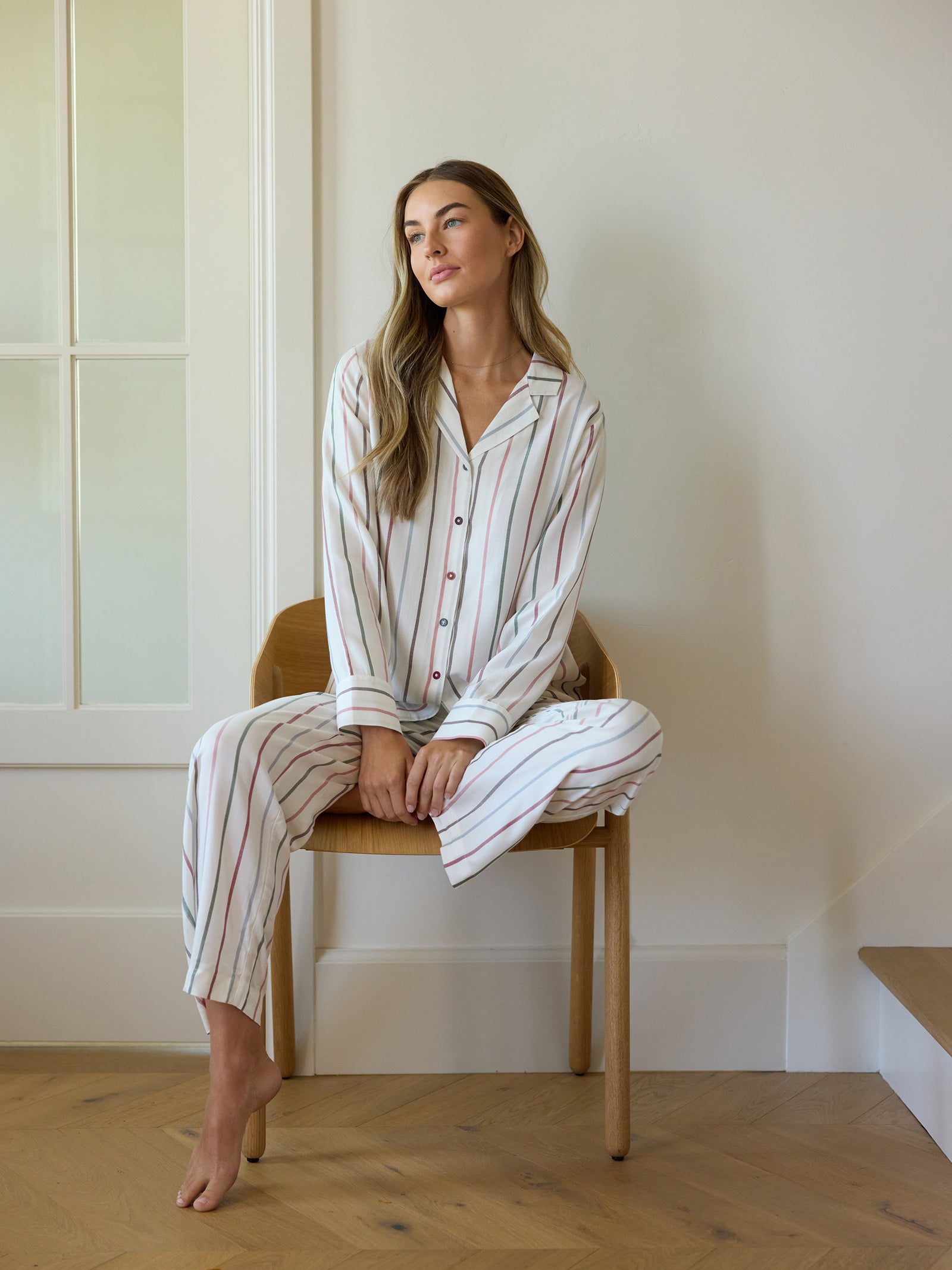 Wearing a Cozy Earth Women's Soft Woven Long Sleeve Pajama Top, a woman with long hair sits relaxed on a wooden chair. The room features light-colored walls and a wood floor, with a window partially visible in the background. 