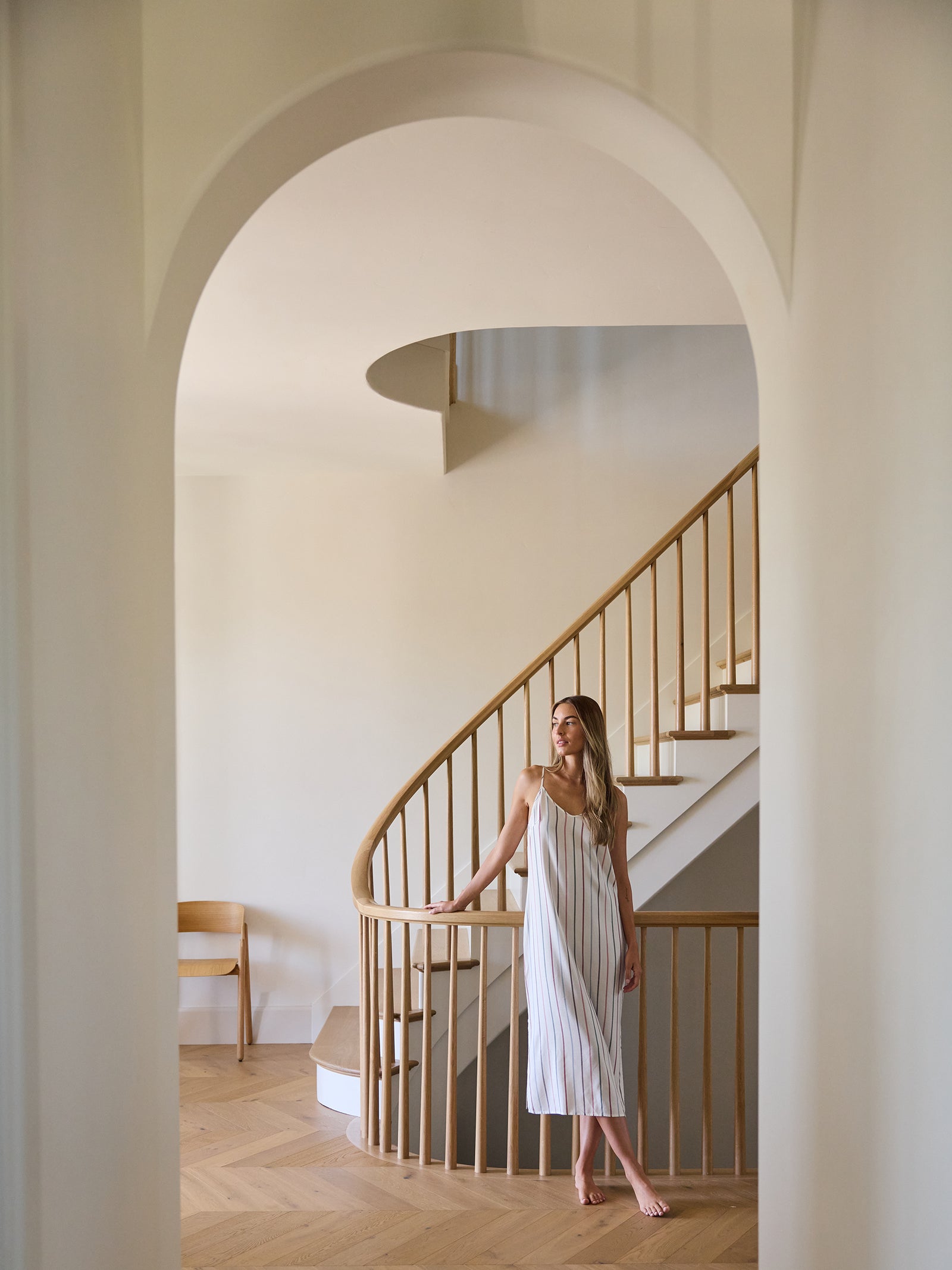 A woman wearing the Women's Soft Woven Nightgown by Cozy Earth stands barefoot on a wooden spiral staircase, framed by an archway. The room has a minimalistic design with light-colored walls and a wooden chair in the background. 