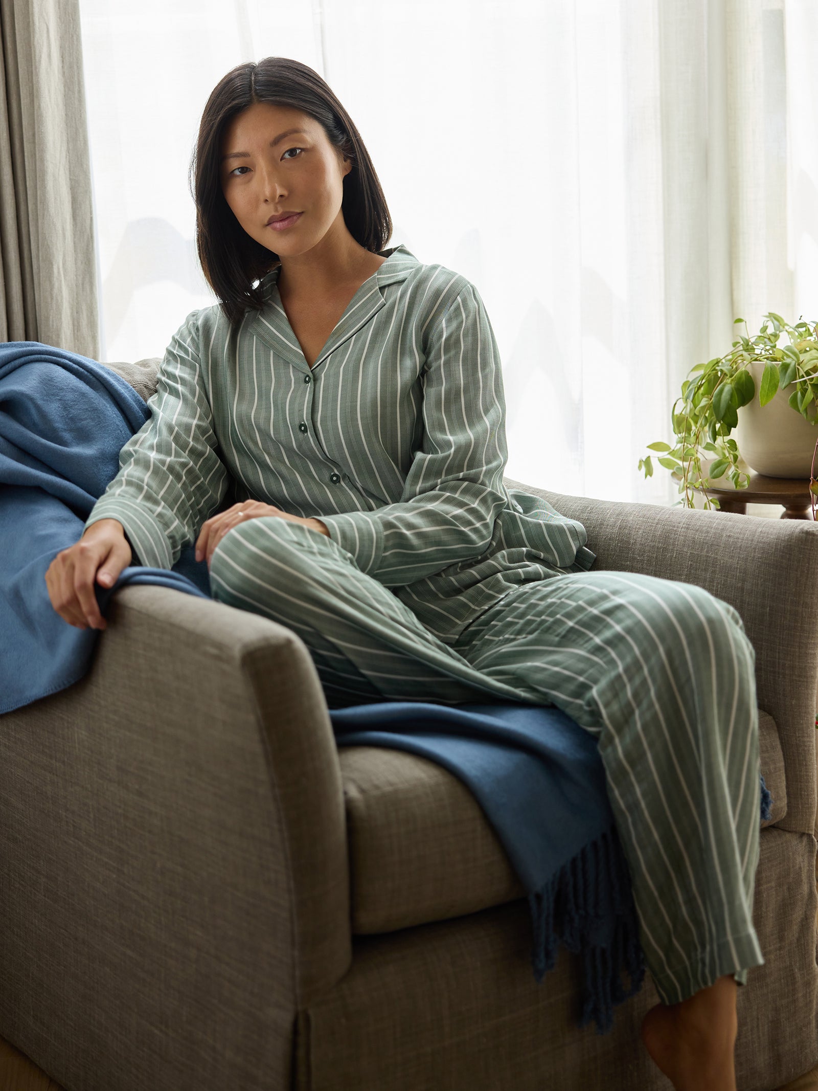 A person with long black hair sits on a sofa wearing Cozy Earth's Women's Soft Woven Pajama Pant. They are leaning against a blue blanket, with a potted plant in the background. The room is softly lit with light filtering through sheer curtains. 
