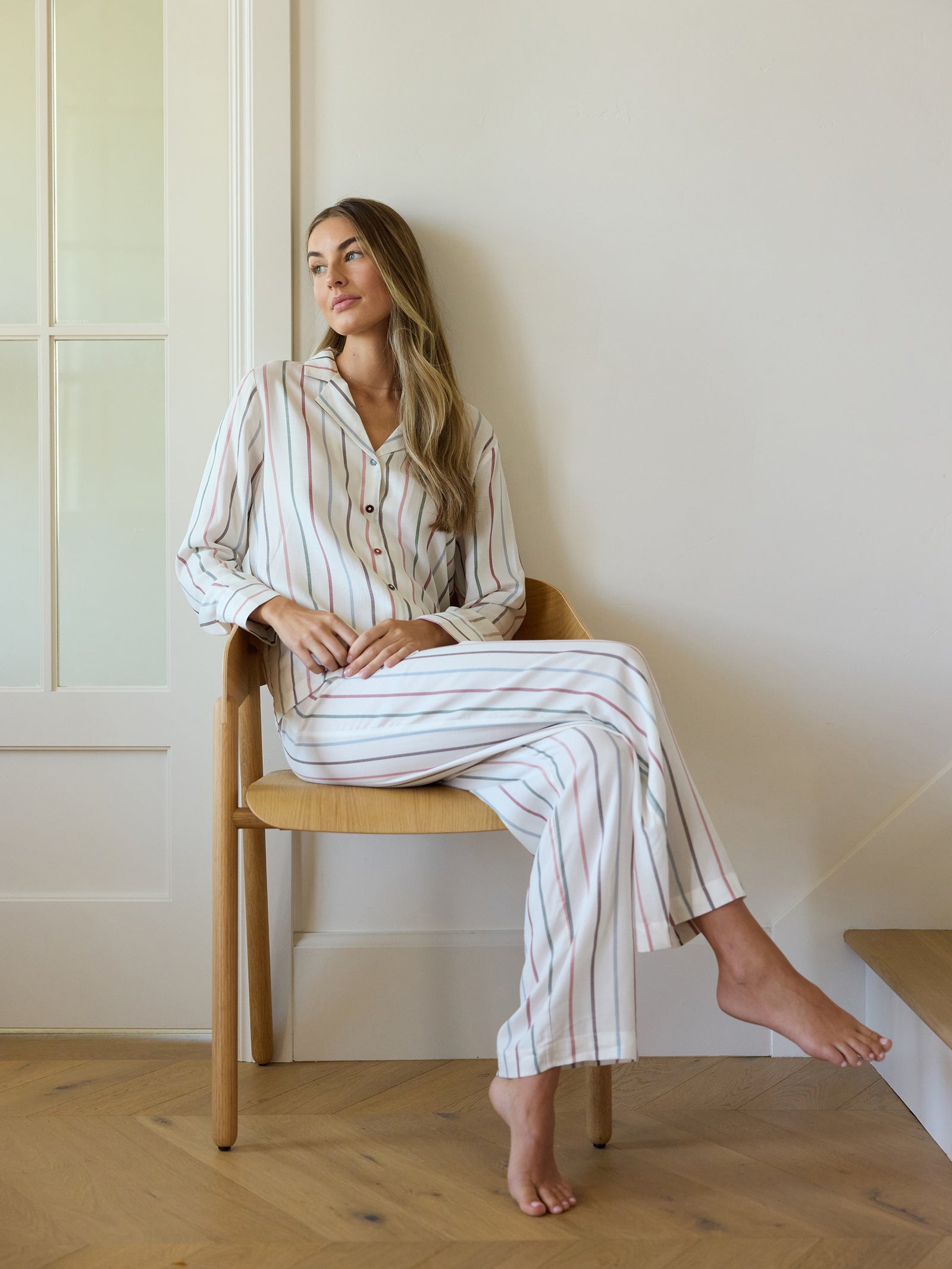 A woman wearing Cozy Earth's Women's Soft Woven Pajama Pant sits cross-legged on a wooden chair in a room with light-colored walls and wooden flooring, gazing out thoughtfully. 