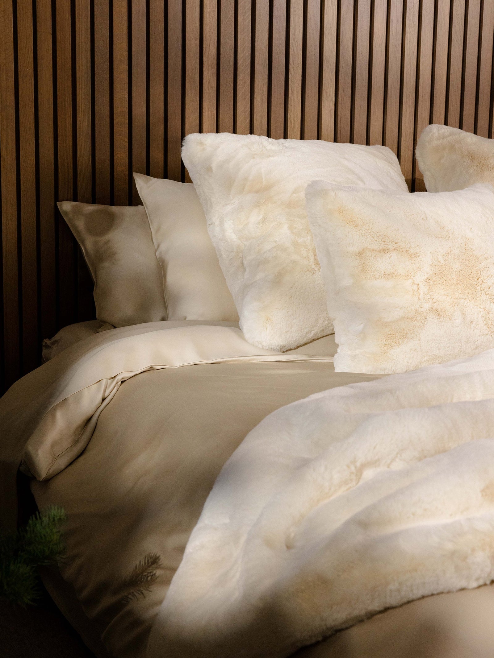 The Cuddle Pillow from Cozy Earth is arranged alongside beige satin bedding and fluffy white pillows on a cozy bed. A soft, matching blanket is draped over it. The backdrop showcases a wooden panel wall. 