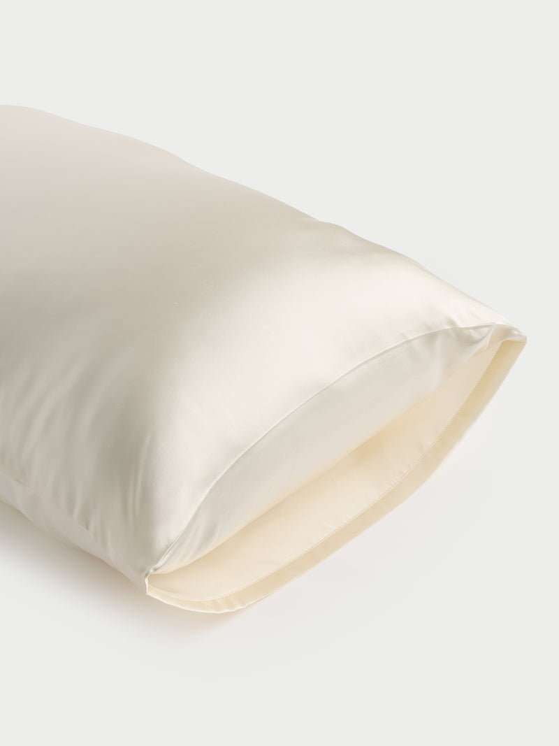 A close-up of a Cozy Earth Silk Pillowcase in a cream color, with one end slightly open. The smooth, shiny fabric softly reflects light, set against a neutral off-white background.