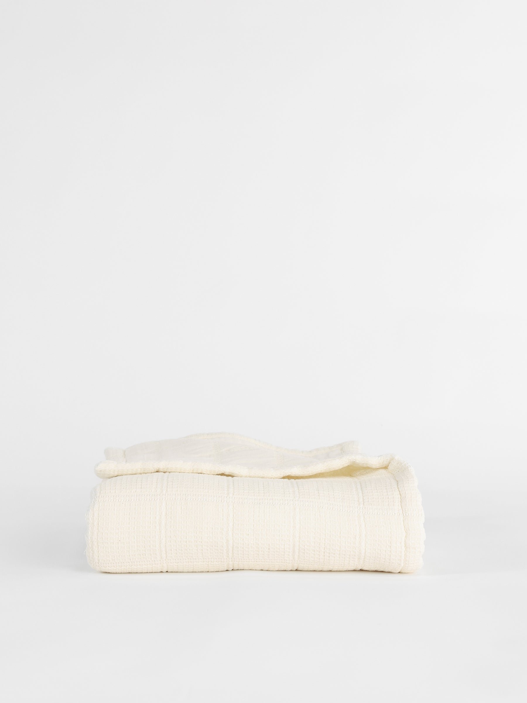 A neatly folded Cozy Earth Waffle Windowpane Blanket, available in beige or off-white and featuring a textured grid-like pattern, is placed against a plain white background. 