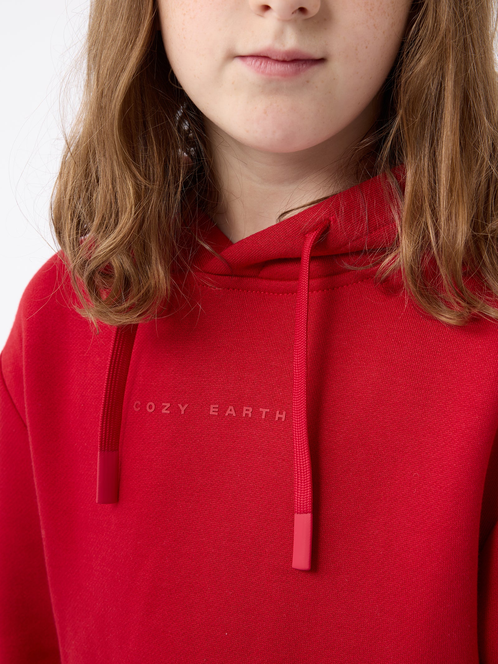 A person with shoulder-length brown hair is wearing the Kid's CityScape Hoodie by Cozy Earth, which is red and has "COZY EARTH" printed on the chest. The background is white. 