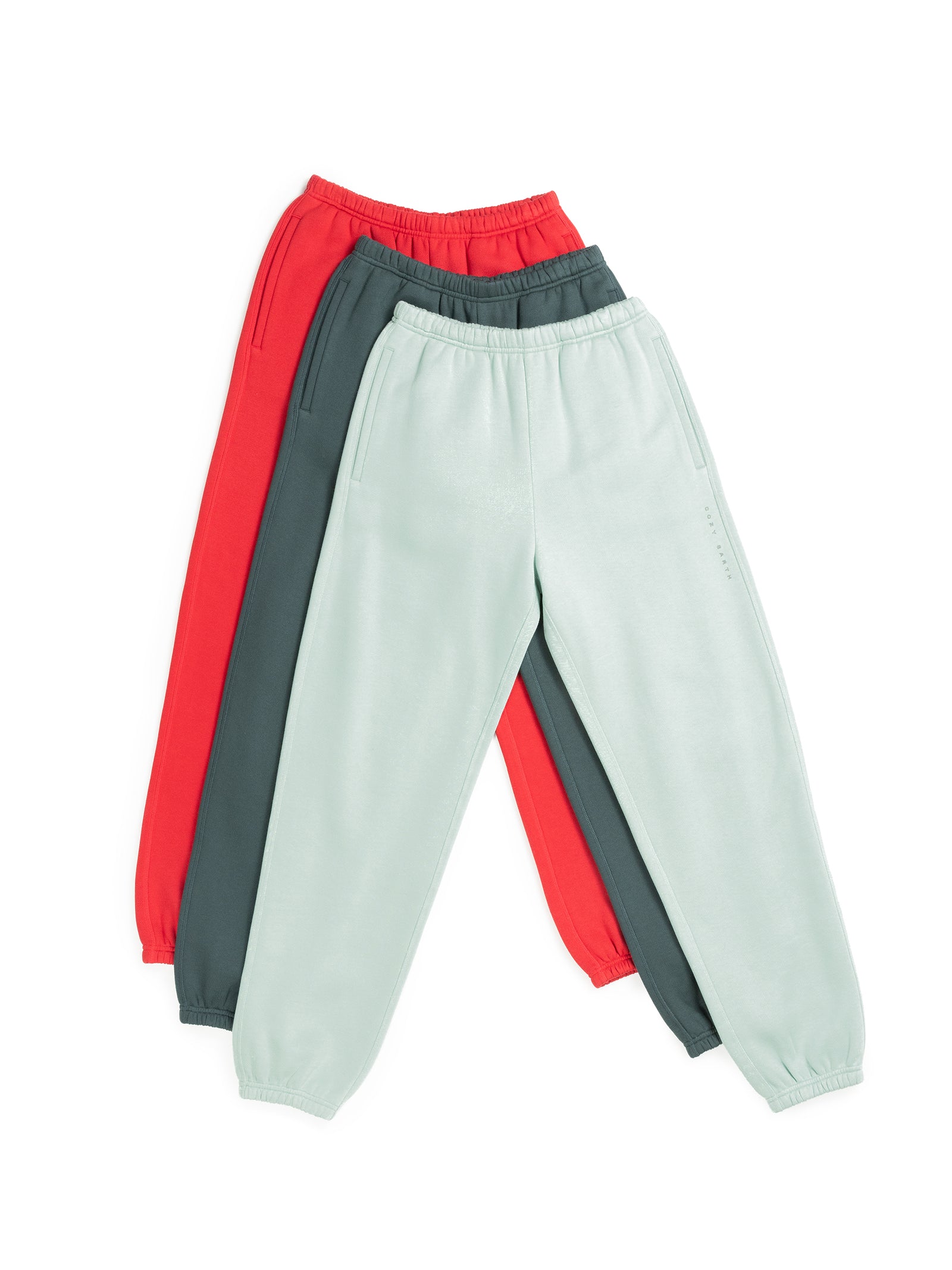 Three pairs of the Women's CityScape Sweatpants by Cozy Earth are laid out overlapping each other. The pants come in red, dark green, and light mint green colors. Each pair features an elastic waistband and cuffs, along with side pockets, all arranged on a white background. 