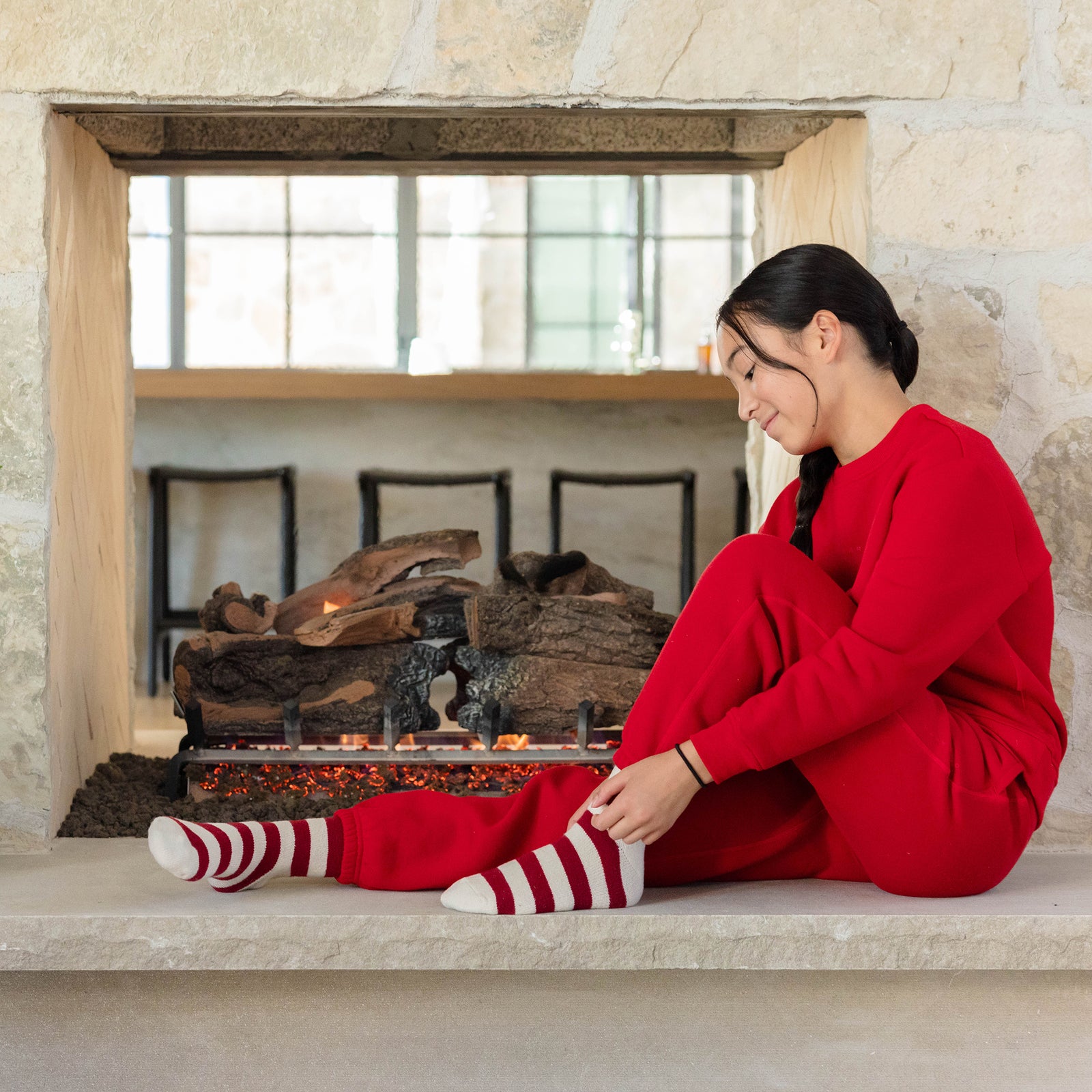 Dressed in Cozy Earth's Kid's CityScape Sweatpants, a person sits on a stone fireplace ledge, adjusting their striped socks. The unlit fireplace stands out in a room filled with bright natural light streaming through large windows in the background. 