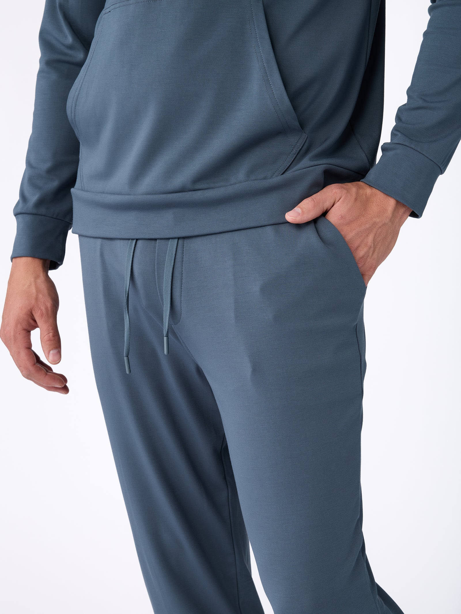 A person is dressed in the Men's Ultra-Soft Bamboo Pullover Crew by Cozy Earth, featuring a blue-gray color. Their right hand is tucked into the pocket of the pants, while the top sports long sleeves and a front pouch pocket against a plain white background. 