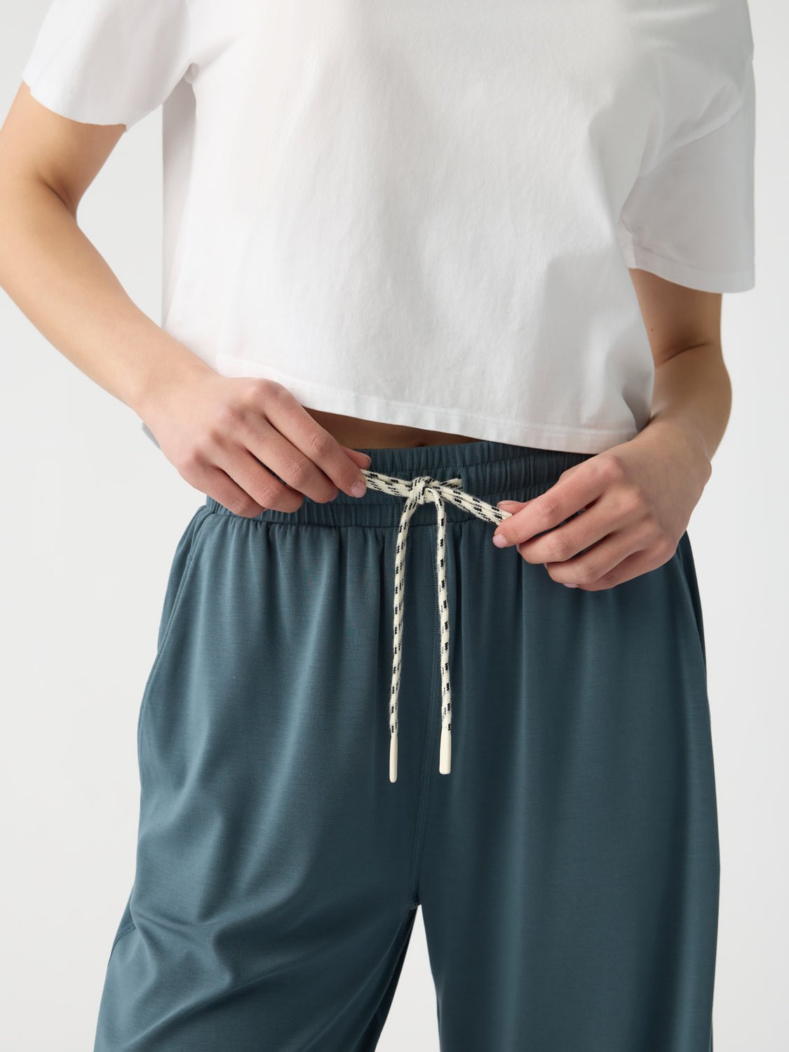 A person in a plain white cropped T-shirt and Cozy Earth's Women's Studio Cropped Wide Leg Pant in teal adjusts the contrasting white and navy drawstring. The casual outfit stands out against a plain backdrop, highlighting the clothing. 