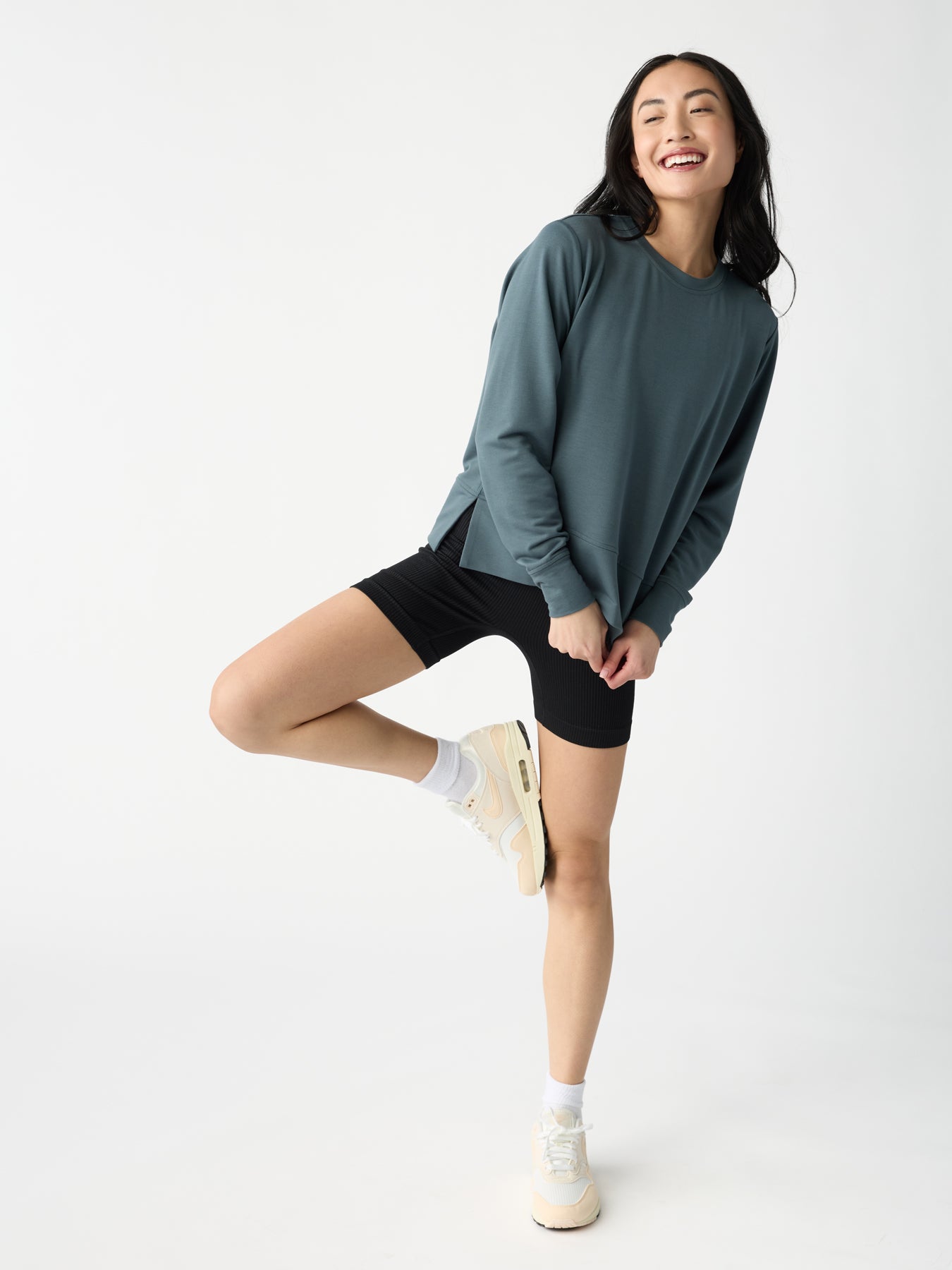 A person smiles with eyes closed, wearing a Cozy Earth Women's StudioLite Crewneck in teal, black shorts, white socks, and cream sneakers. They balance on one leg against a plain white background. 