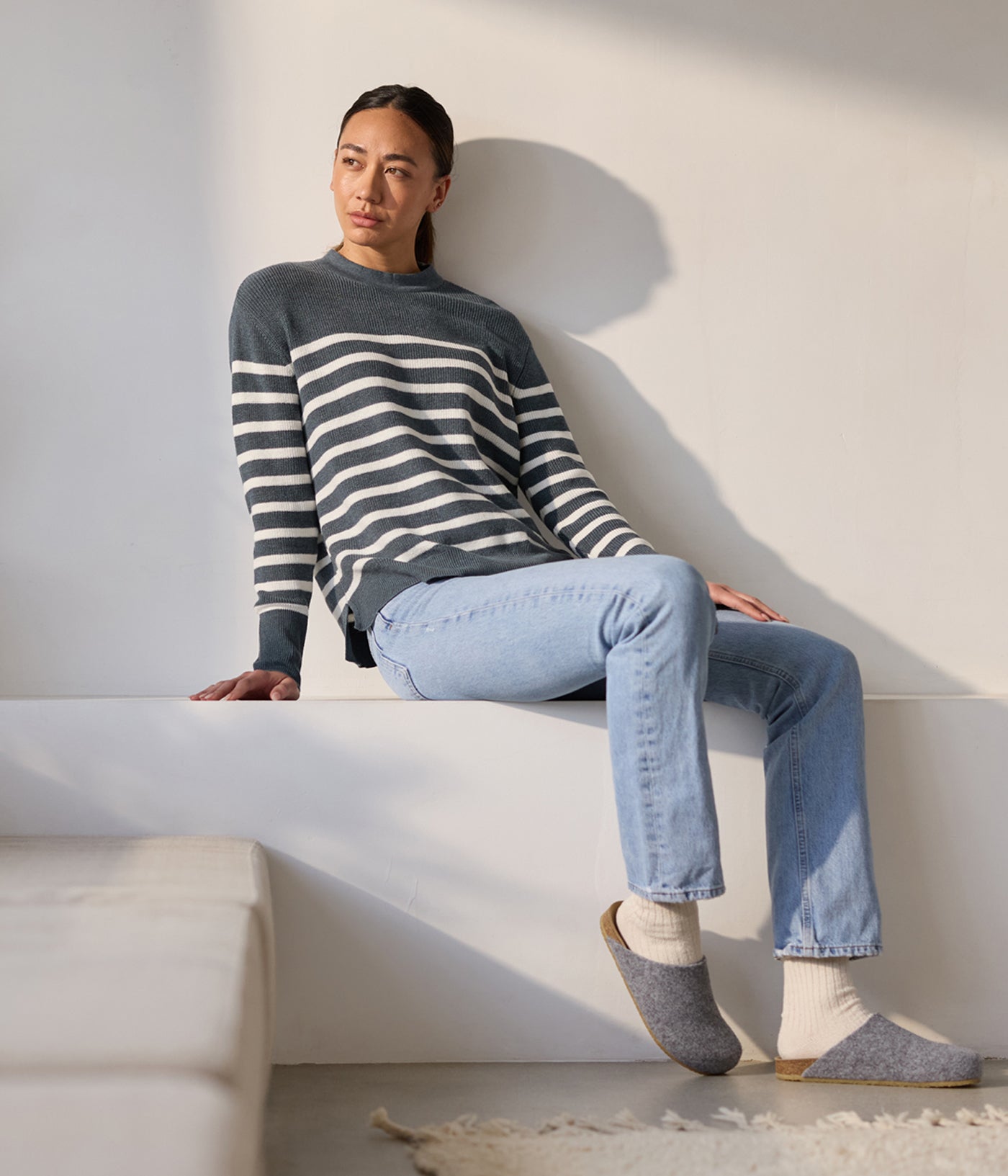 A person sits on a ledge in a dark striped Women's Rowan Sweater by Cozy Earth, paired with light blue jeans and gray slippers over white socks. Sunlight fills the room, casting soft shadows. 