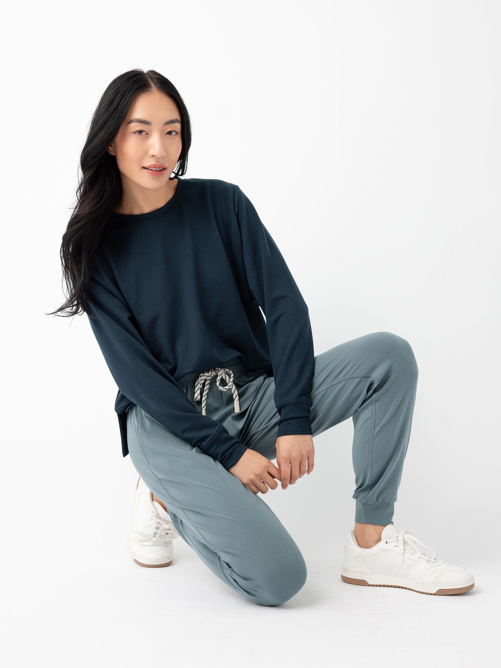 A person with long black hair is posing on one knee against a white background. They are wearing a dark blue long-sleeved shirt, Cozy Earth's Women's Studio Jogger in light blue with a drawstring, and white sneakers. 