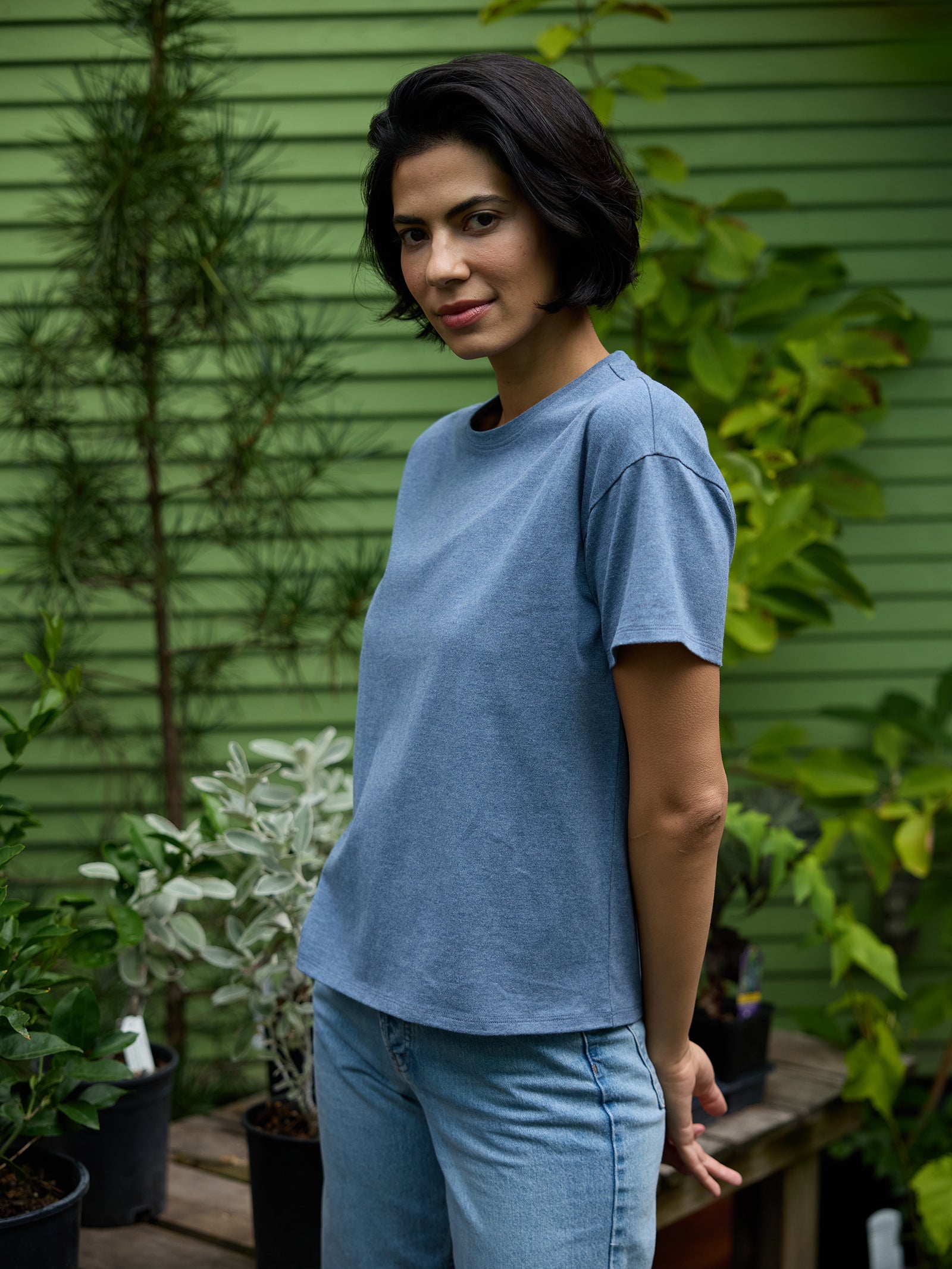 A person with short dark hair stands in a garden wearing a Cozy Earth Women's All Day Tee in blue and blue jeans. They are surrounded by green plants and appear to be in an outdoor setting with a green wooden wall in the background. 