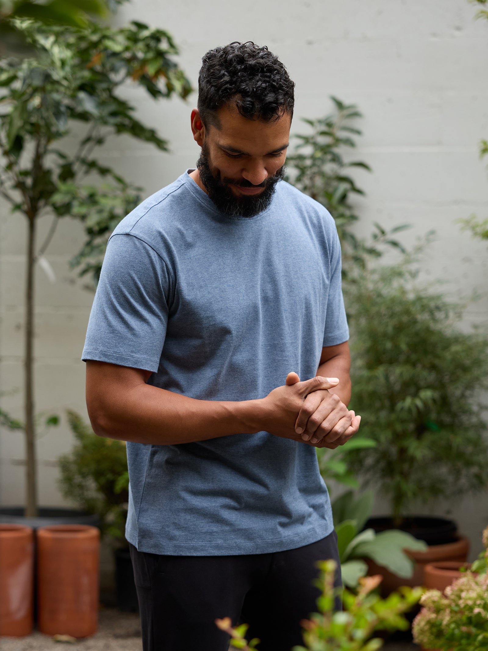 A bearded man with short curly hair stands outdoors in a garden, looking down and smiling. He is wearing the light blue Men's All Day Tee from Cozy Earth paired with dark pants. Potted plants and trees in the background create a lush, green environment. 