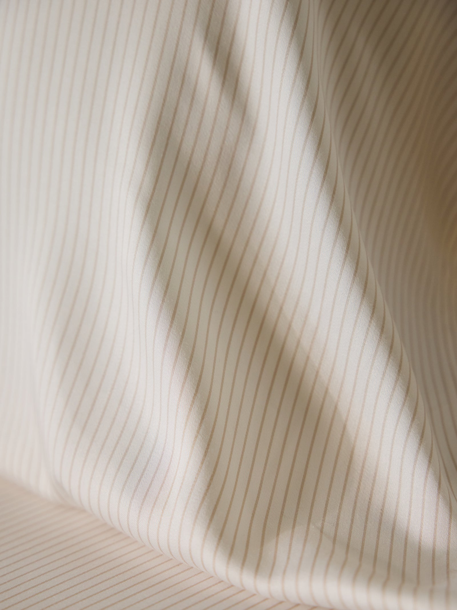 Close-up of a Bamboo Pillowcase by Cozy Earth, showcasing a white fabric adorned with narrow, vertical beige stripes. The fabric is softly draped, creating gentle folds and shadows that enhance its texture and pattern. 