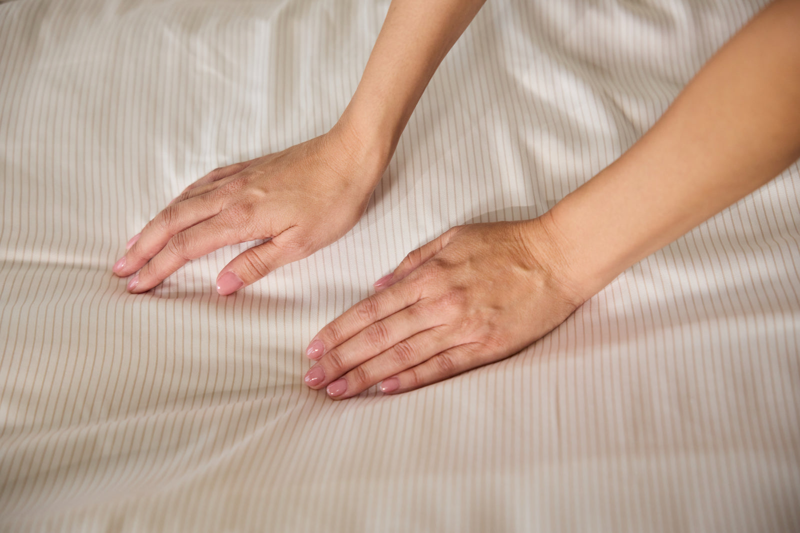 Two hands with neatly trimmed nails are gently pressing down on a smooth, white bamboo fitted sheet from Cozy Earth, creating gentle folds. 