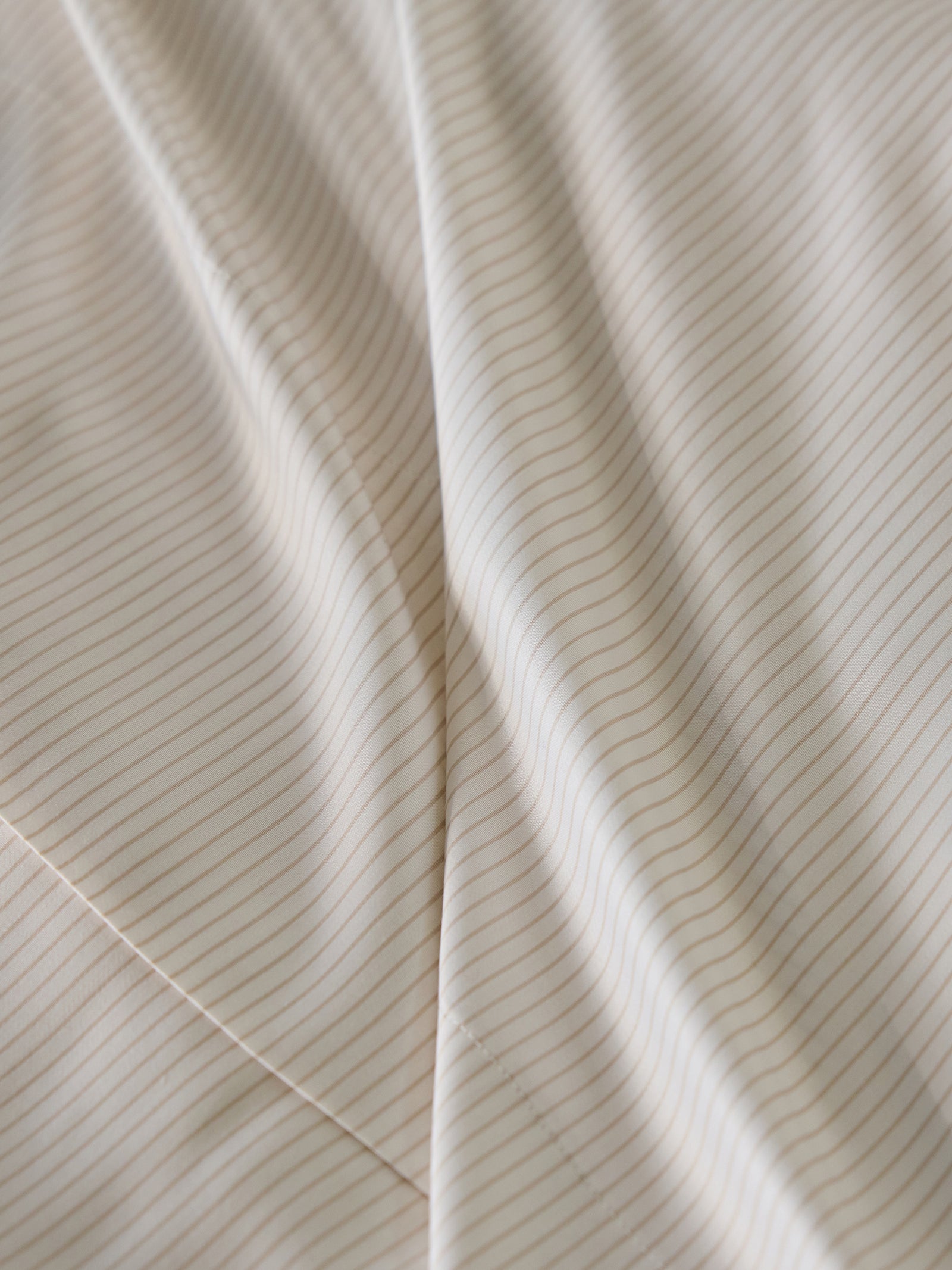 Close-up of Cozy Earth's Bamboo Sheet Set features a soft, cream-colored fabric with thin, brown pinstripes running diagonally. The fabric looks smooth and slightly wavy, creating gentle folds. 