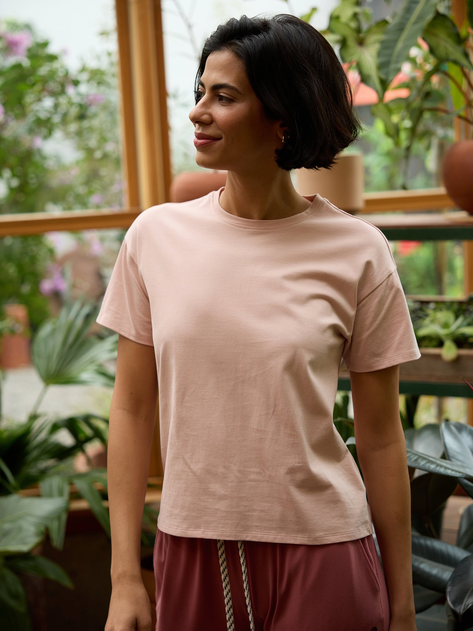 A person with short dark hair, wearing Cozy Earth's Women's All Day Tee in light pink and maroon pants, stands indoors in front of large plants and potted greenery. They are smiling and looking off to the side, suggesting the setting is a greenhouse or indoor garden. 