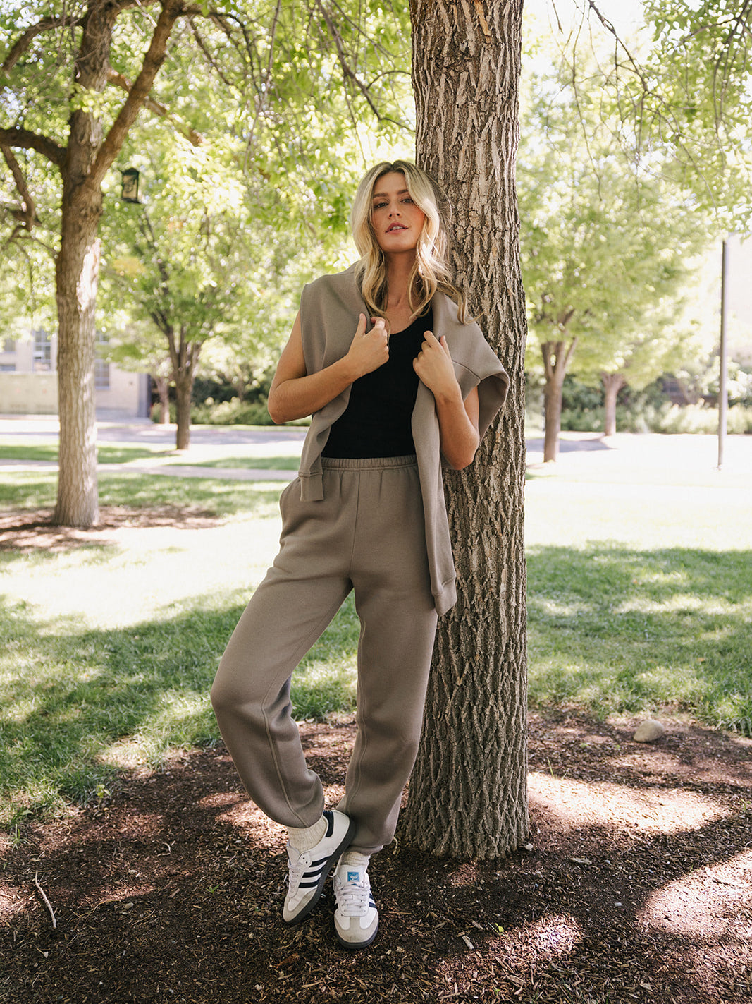 A woman with long blonde hair leans against a tree in a park, dressed casually in Cozy Earth's Women's CityScape Crewneck paired with a beige cardigan and matching beige joggers. She completes her outfit with white sneakers, standing amidst greenery and sunlight filtering through the trees. 