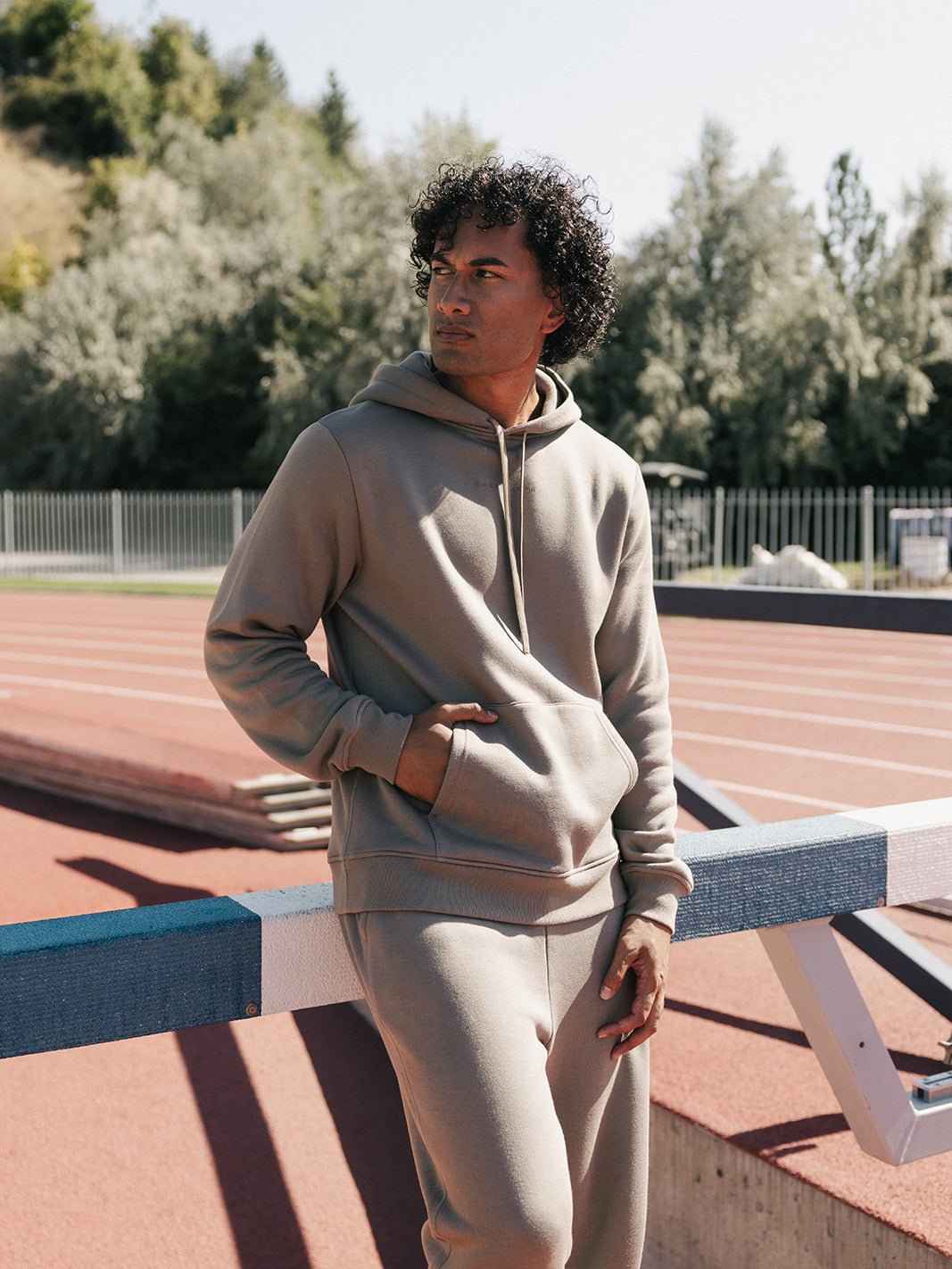 A person with curly hair wearing a beige Men's CityScape Hoodie and matching sweatpants from Cozy Earth stands near a hurdle on an outdoor track field. They have one hand in their pocket and gaze thoughtfully into the distance, with trees and a fence visible in the background. 