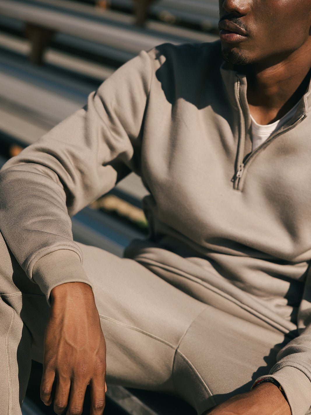 A person wearing a Cozy Earth Men's CityScape Quarter Zip in light brown, along with matching pants, is seated outdoors. The image focuses on the upper body and part of the legs, with the person's face partially visible and cropped out of the frame. The background consists of outdoor bleachers. 