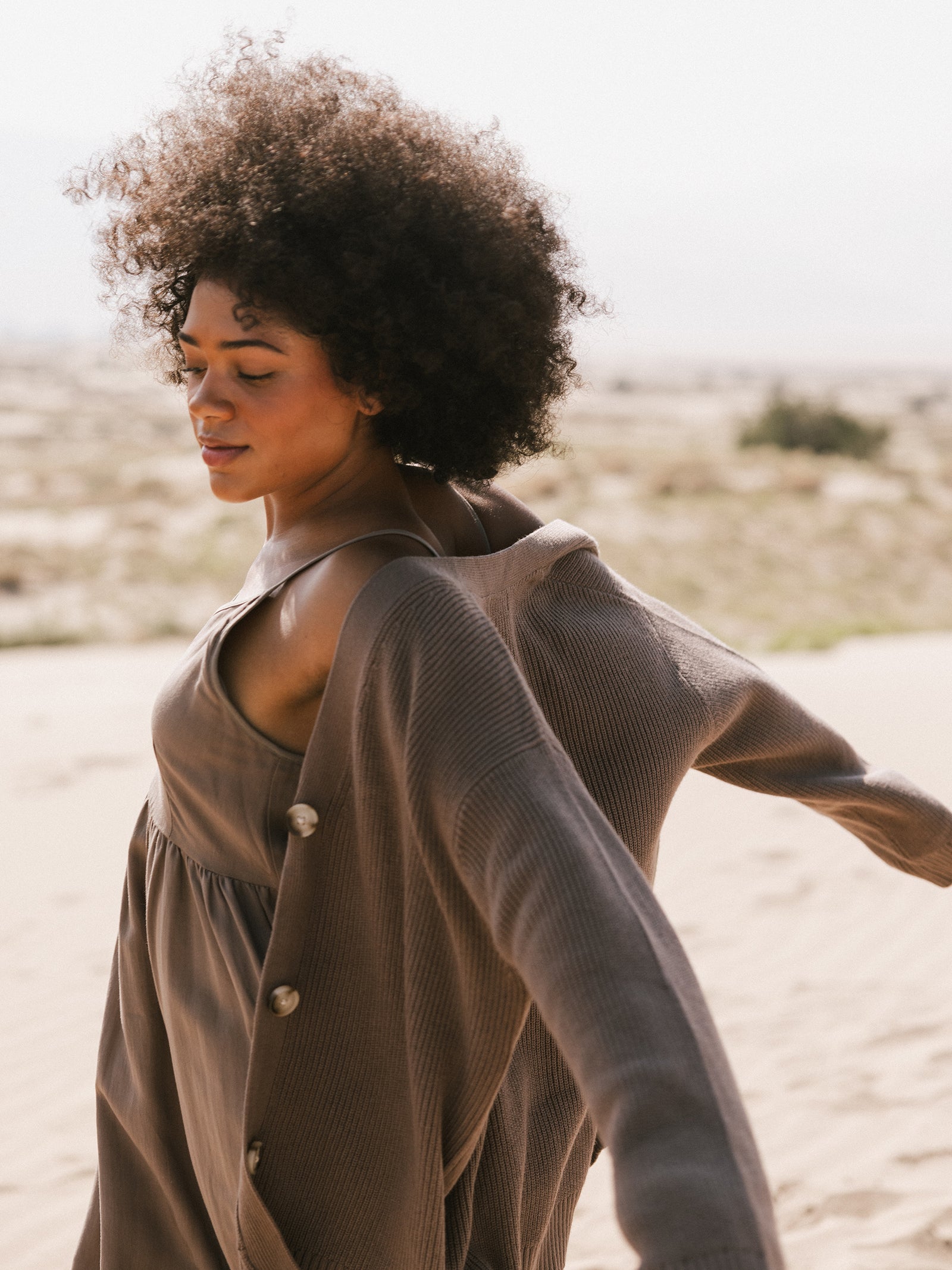 A woman with curly hair stands on a sandy landscape, wearing a loose-fitting dress and the Oversized Classic Cardigan by Cozy Earth. She appears serene with closed eyes, as her hair and clothes drift slightly in the breeze. 